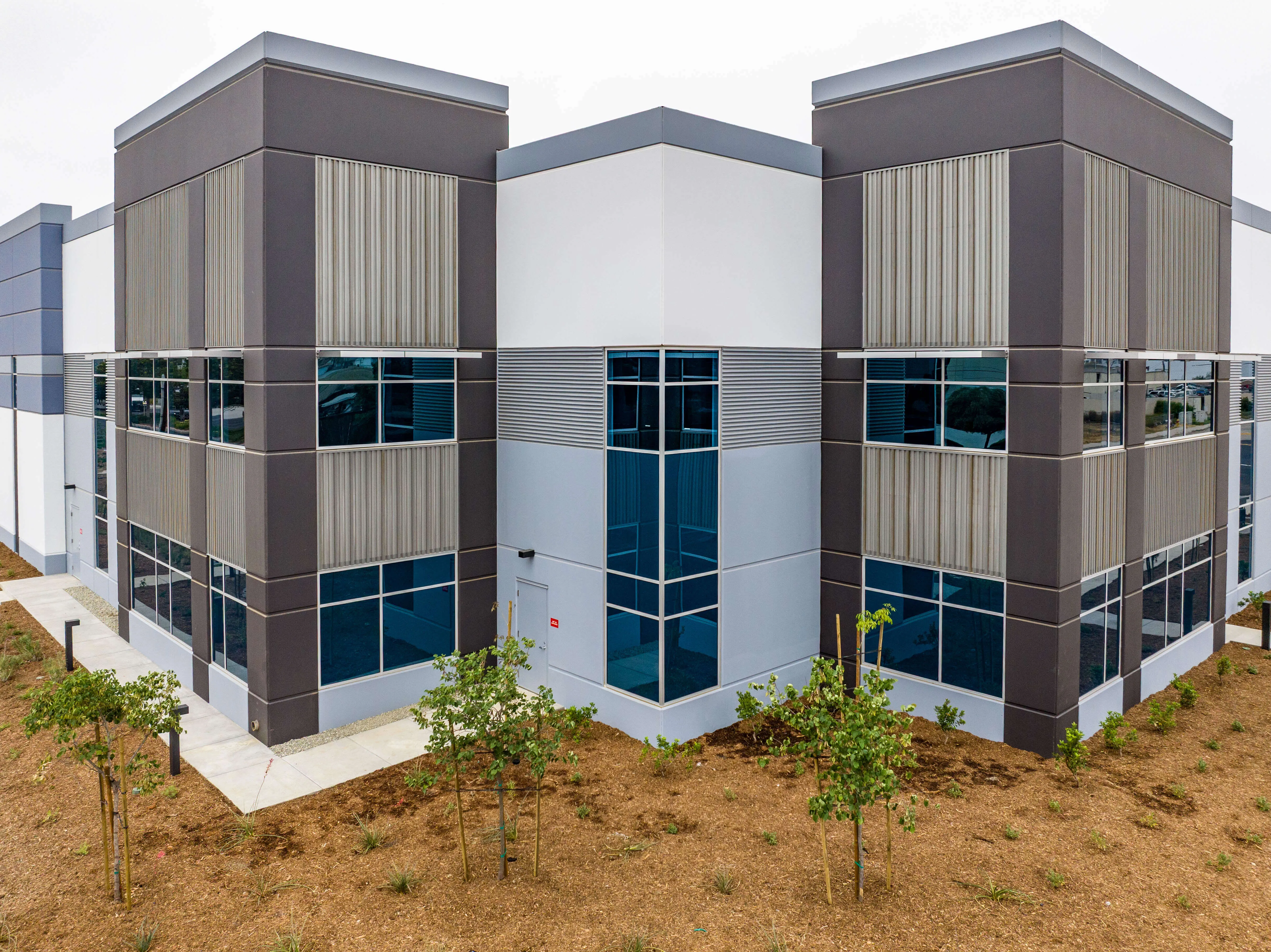 Modern commercial building with large blue-tinted windows and grey and white exterior walls, surrounded by young landscaping with small trees and mulch.