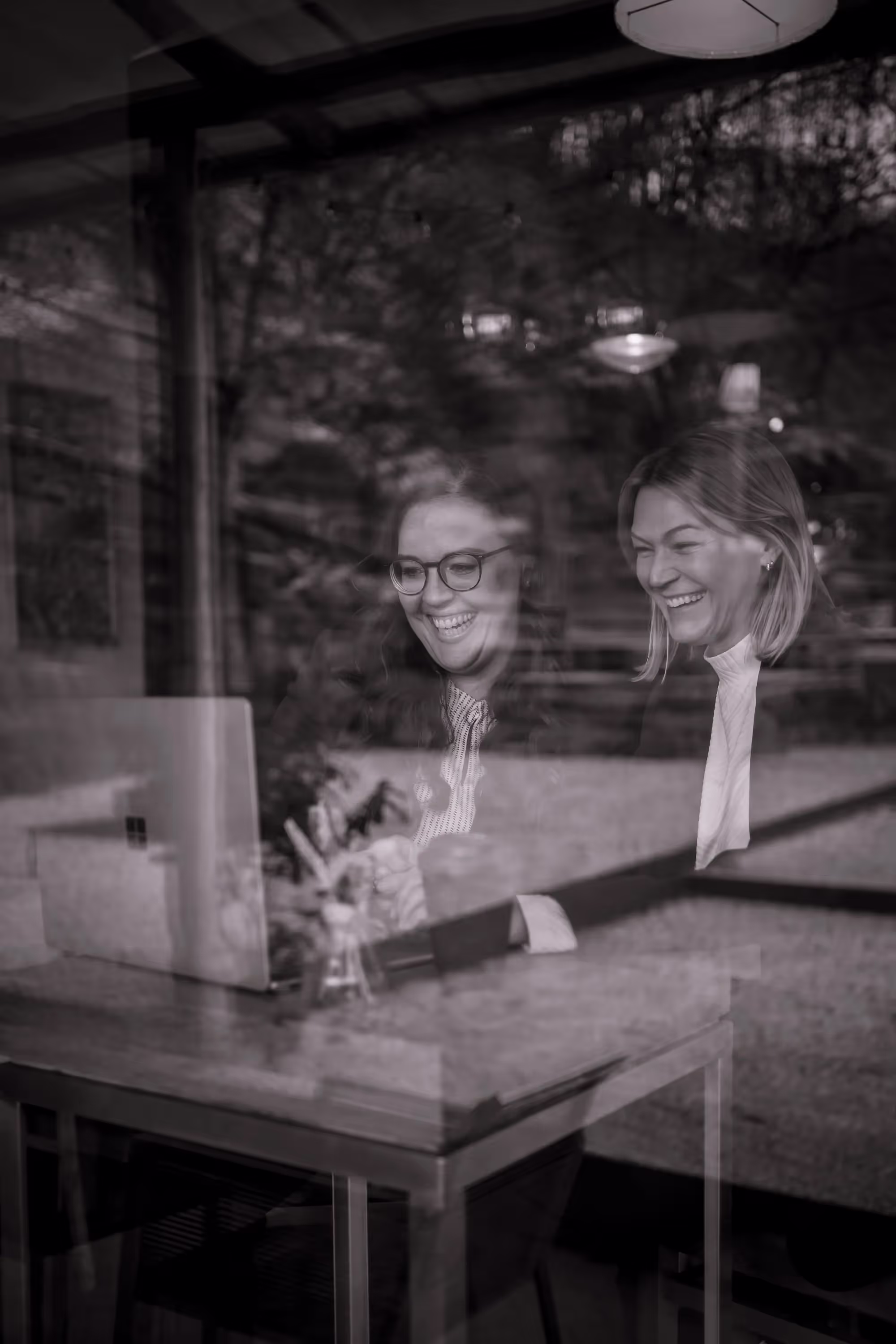 Zwei lachende Frauen bei der Arbeit am Laptop, durch ein Fenster aufgenommen – authentischer Moment im Stil moderner Corporate Fotografie