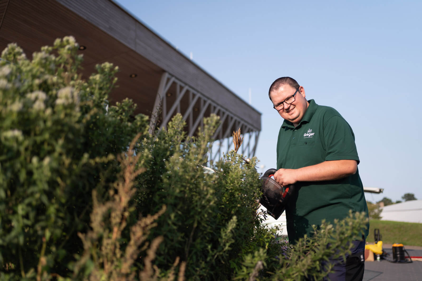 Ein Mitarbeiter der Firma Greger Haus und Garten GmbH schneidet Büsche vor einem modernen Firmengebäude an einem sonnigen Tag.