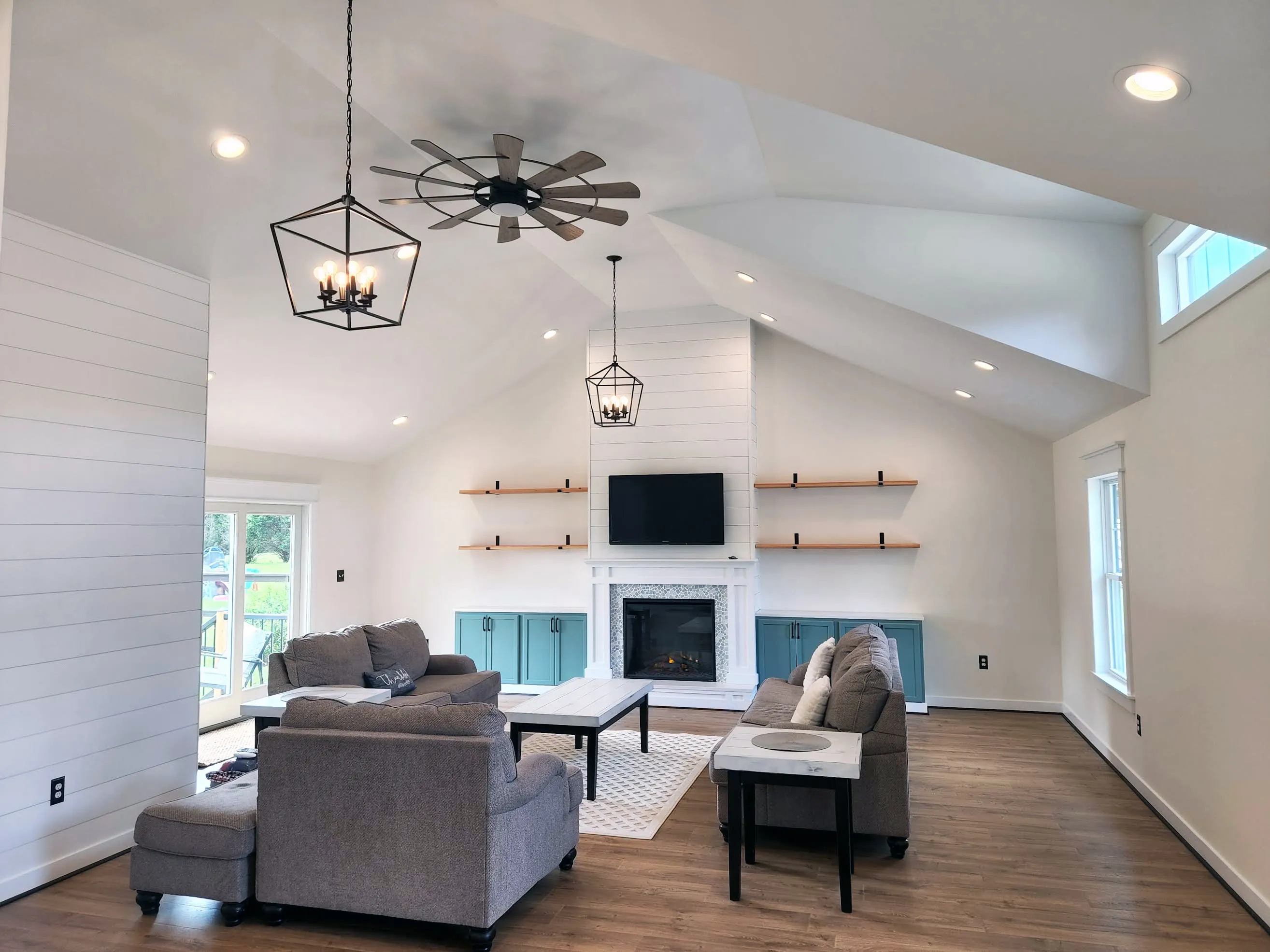 Modern living room with vaulted ceiling, gray sofas, a fireplace, and teal cabinets under floating shelves.