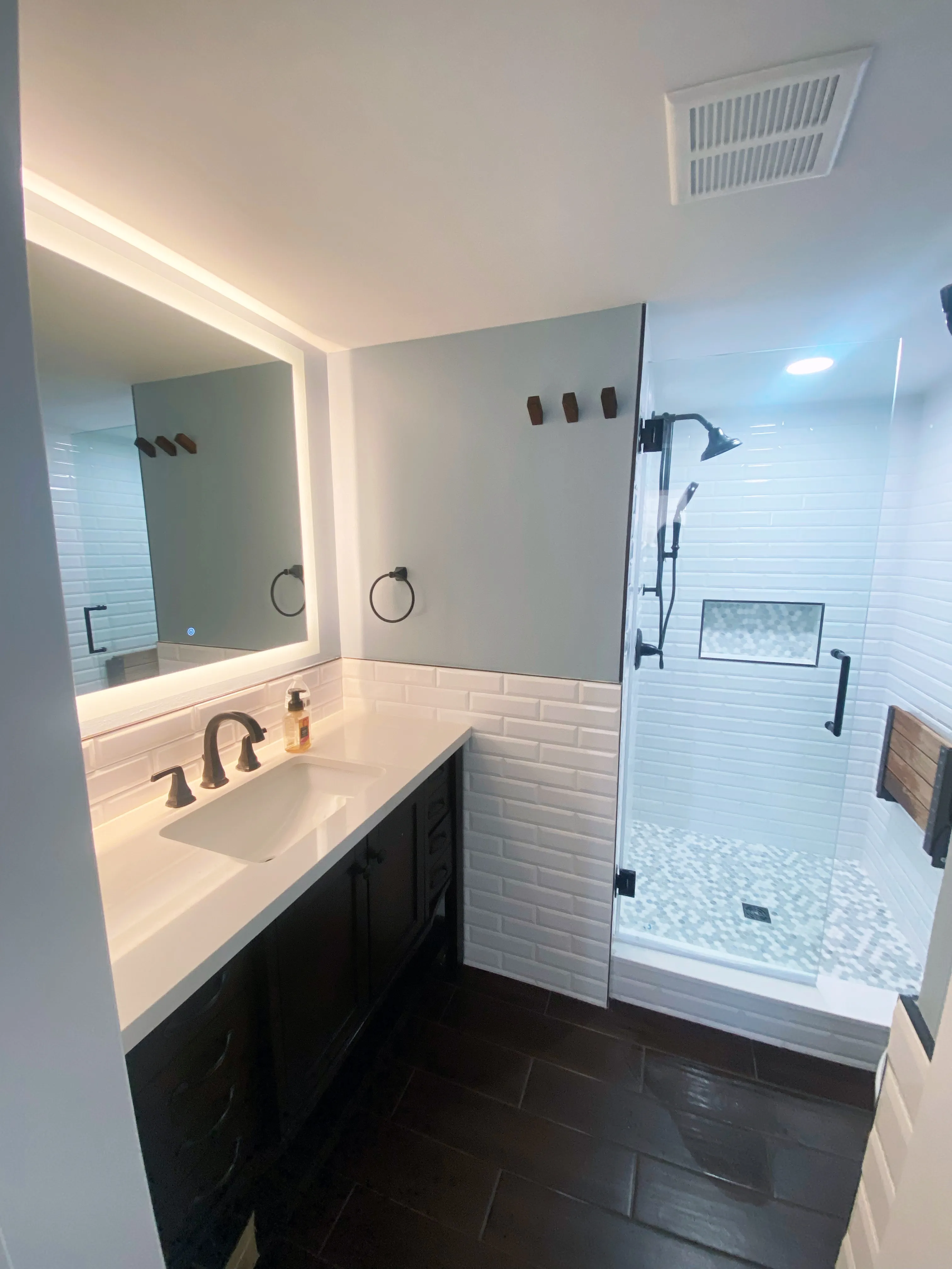 Modern bathroom with dark wood vanity, white countertop, backlit mirror, and glass shower with patterned tile floor.