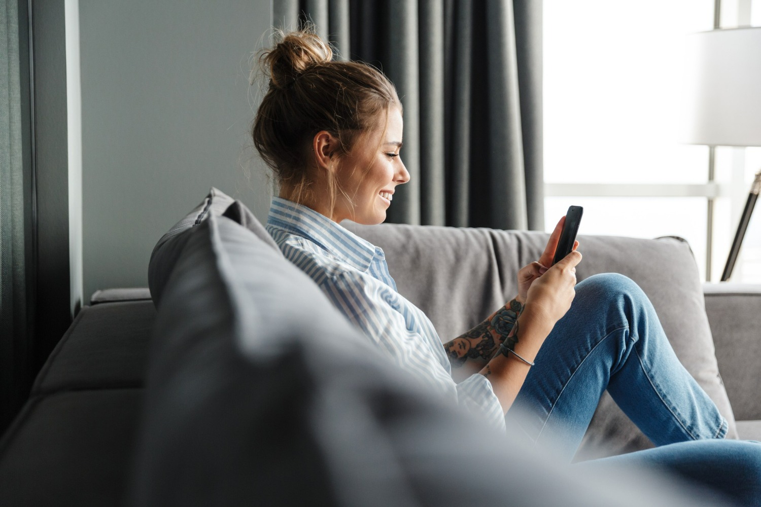 Young woman sitting on a sofa and smiling while using her smartphone, illustrating user-friendly mobile access to a HIPAA-compliant alumni app