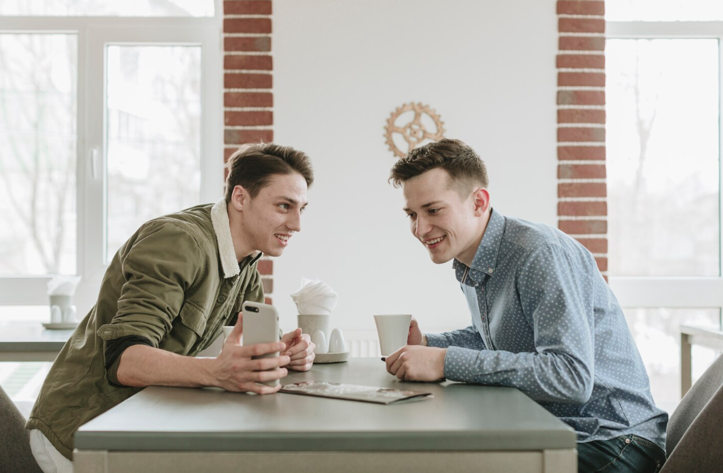 Two men sit at a table in a bright café, smiling and using a smartphone while exploring addiction recovery app development in a casual, friendly setting