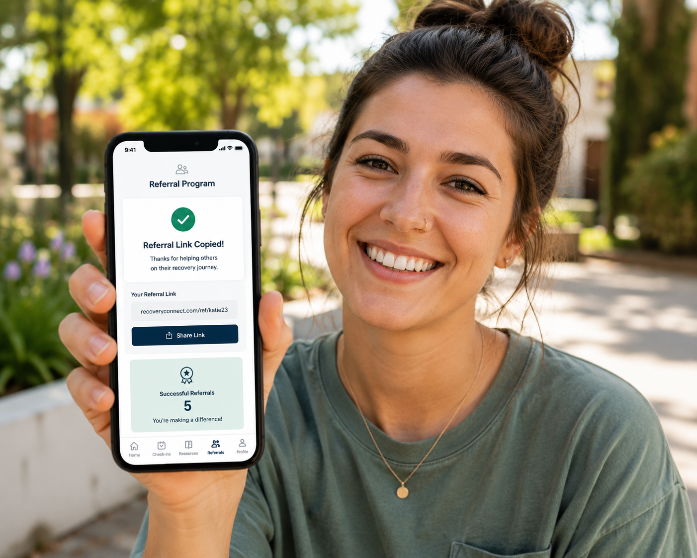 Smiling recovery alumni holding a smartphone displaying a behavioral health referral program with a referral link and confirmation screen, representing alumni referral tracking rehab and digitally connected alumni engagement