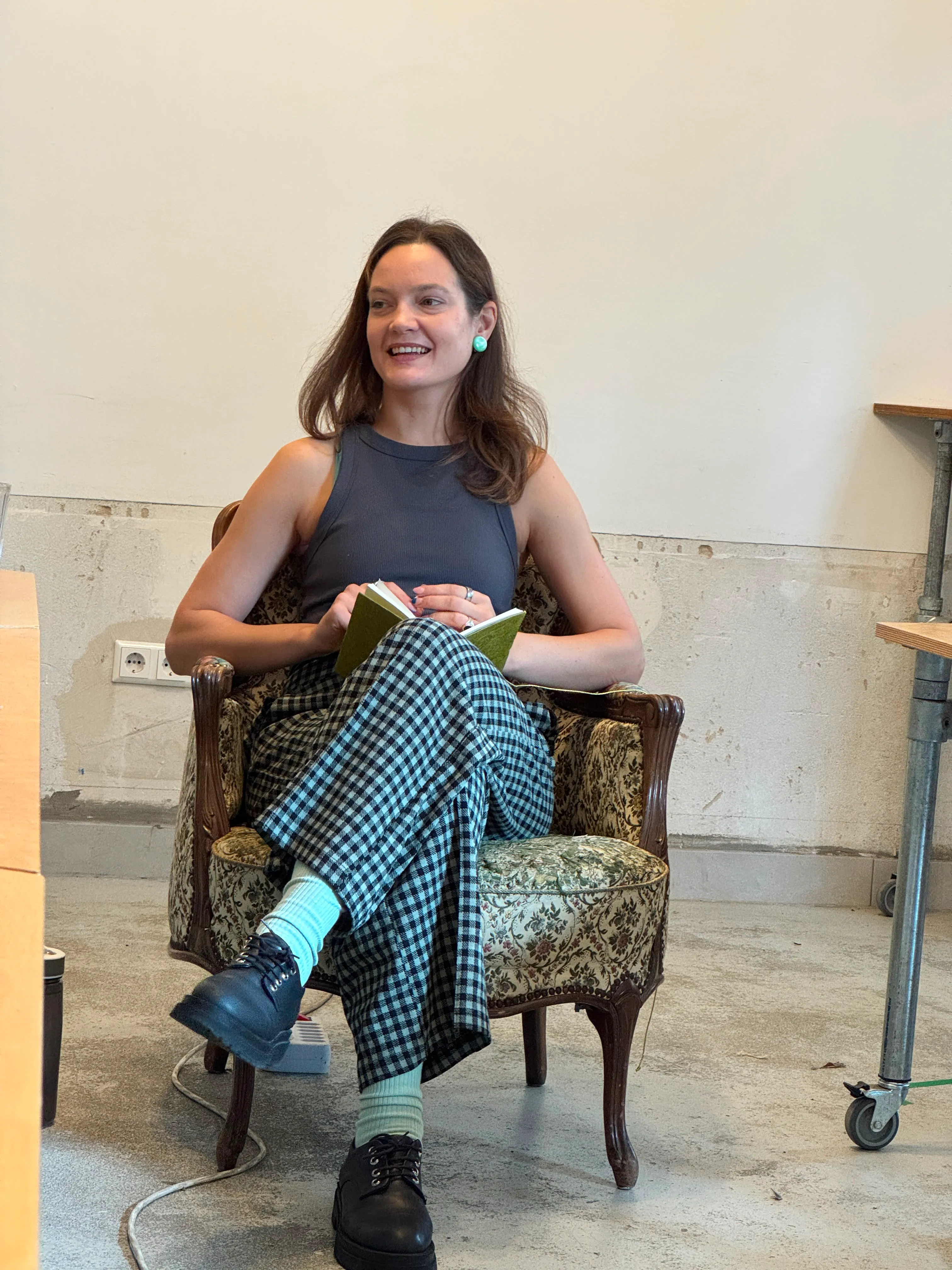 Smiling woman sitting on vintage armchair holding a book, wearing checkered pants and black shoes.
