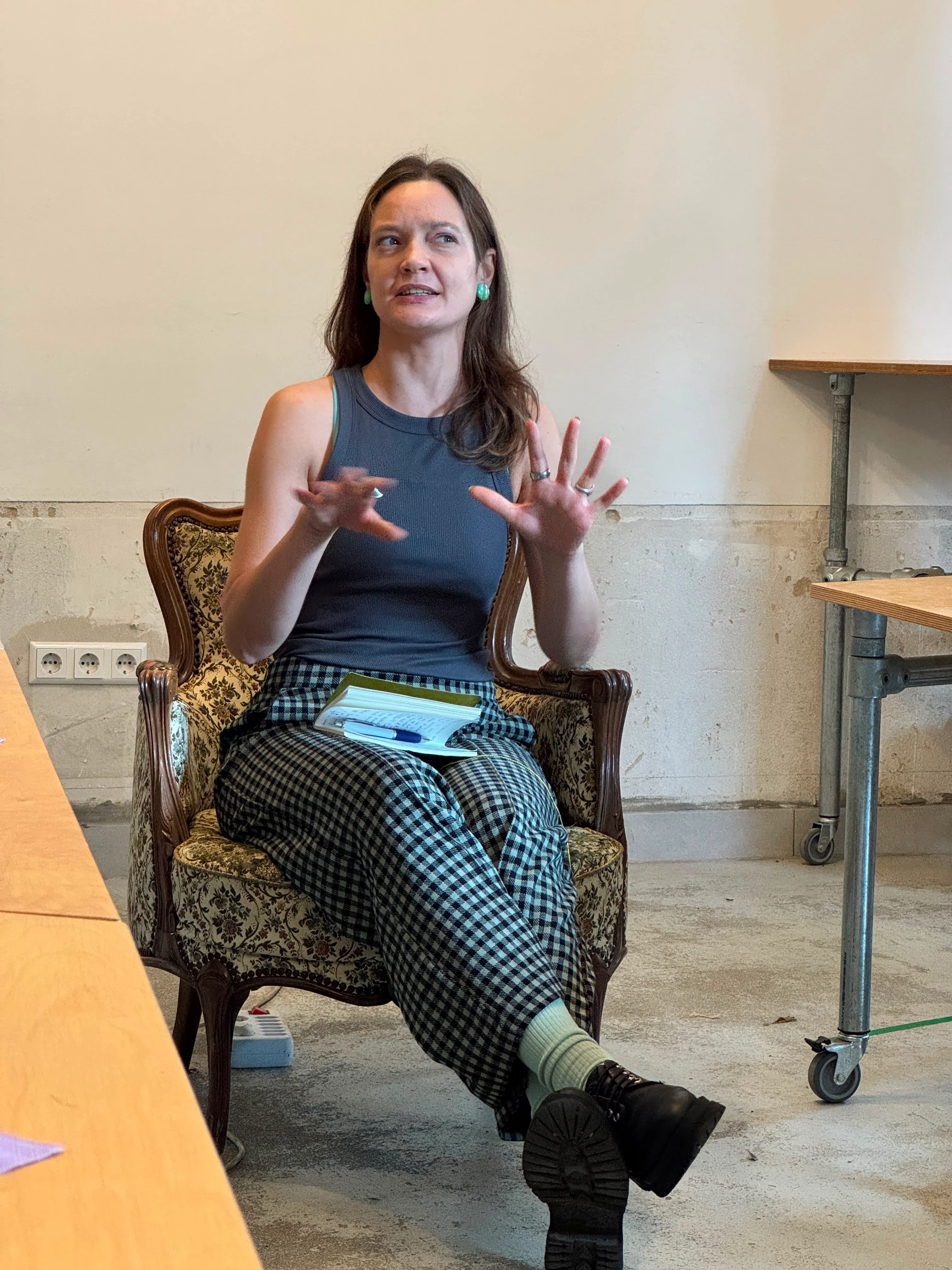 Woman seated on ornate chair gestures with hands while holding notebook and pen on lap.