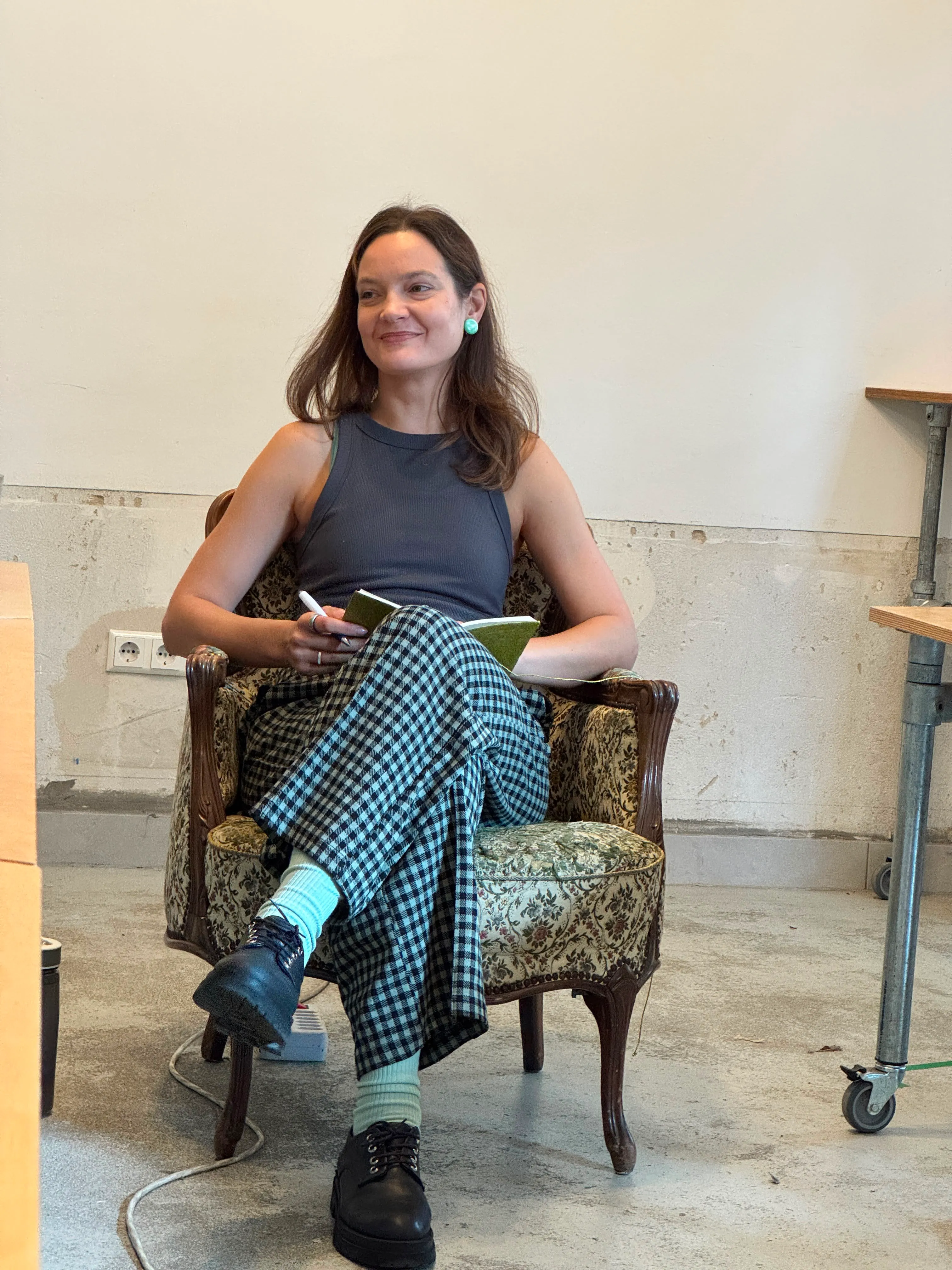 A woman with long brown hair sits cross-legged on a vintage floral armchair, wearing a sleeveless dark gray top, checkered wide-leg pants, mint green socks, and black shoes, holding a green notebook and pen indoors with a plain wall behind her.