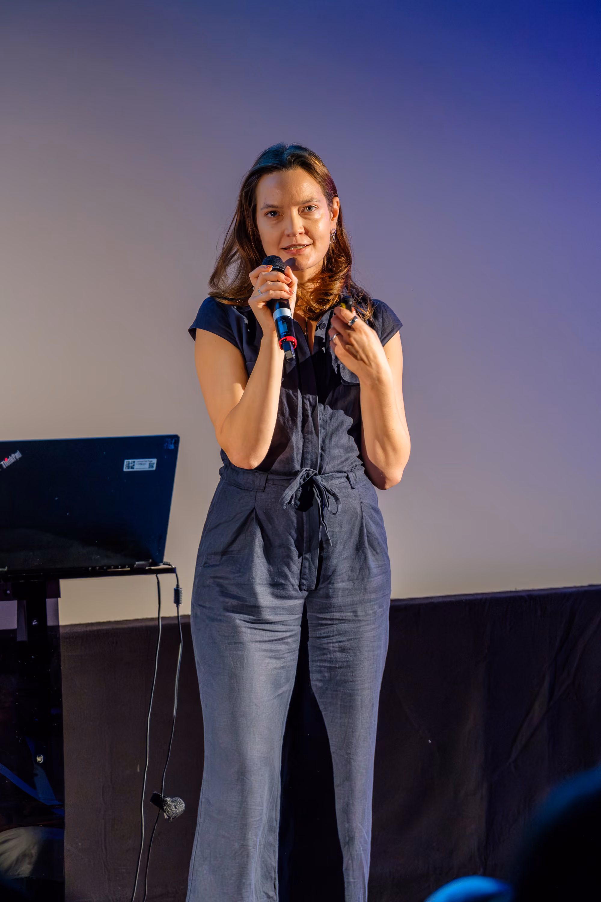 A woman stands on stage holding a microphone, speaking to an audience, wearing a dark jumpsuit.