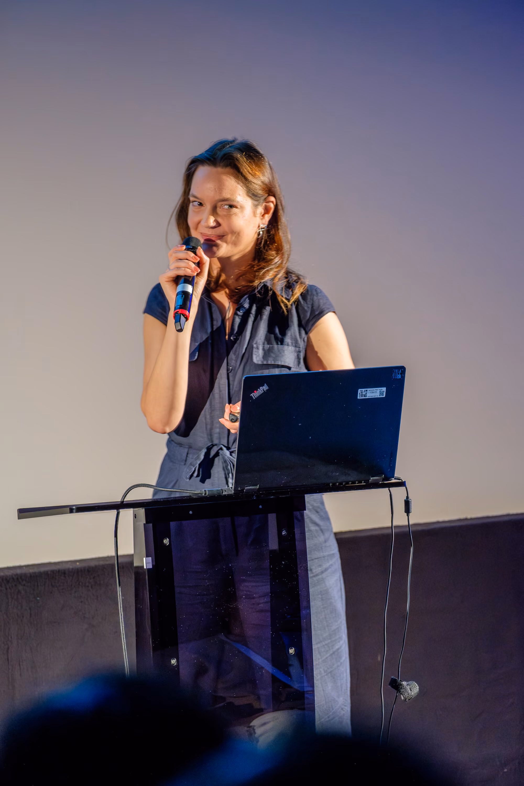Magda - a woman with long brown hair wearing a dark sleeveless dress is speaking at Freelance Unlocked conference into a microphone while standing behind a clear podium with a laptop on it, against a plain light background.