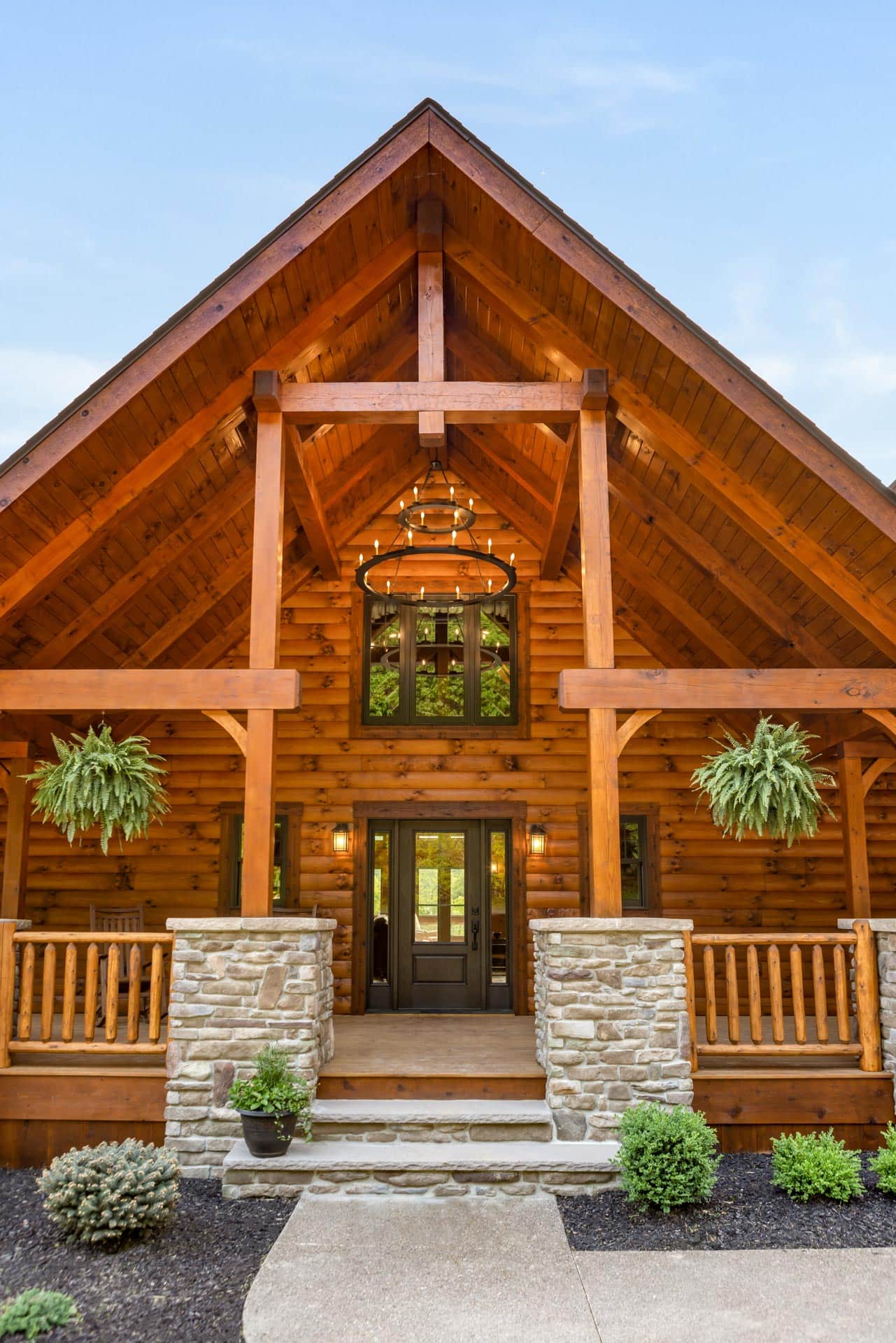 The exterior of a log home featuring a stone base and wooden beams, with a large front porch adorned with hanging plants and a circular light fixture. This custom home showcases traditional log cabin architecture.
