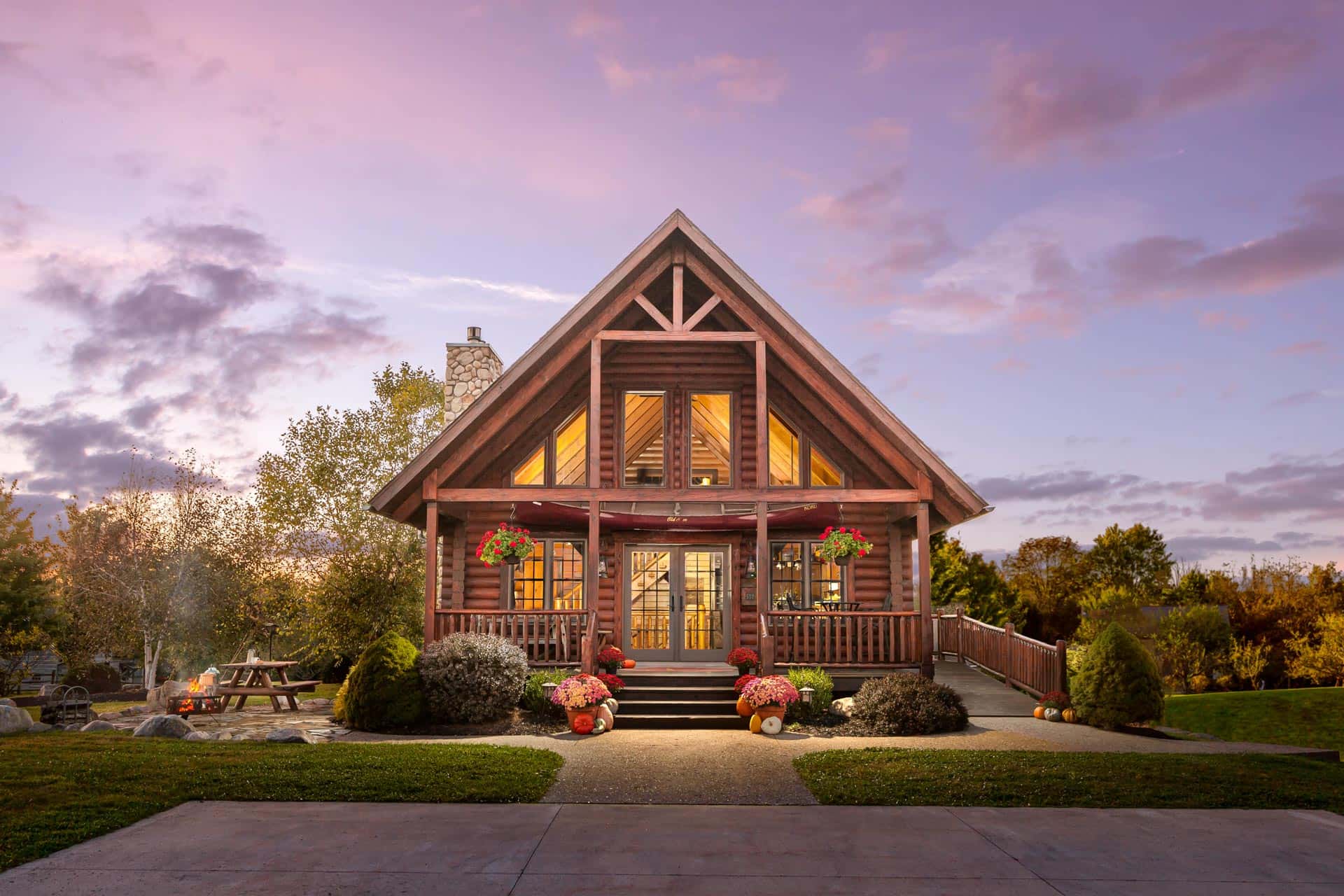 A beautifully designed log cabin with a steep roof and large front porch, surrounded by trees and a landscaped garden. The exterior features wood siding and large windows, with decorative flower arrangements on the porch. Evening light casts a warm glow.