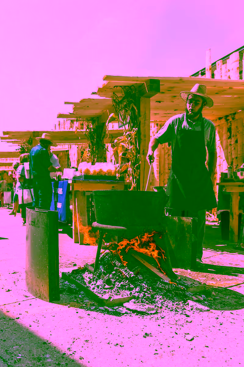 A man in a straw hat and apron stirring a pot over an open flame at a market. Wooden stalls are set up in the background with various products visible. This scene captures the essence of outdoor cooking and community gatherings.
