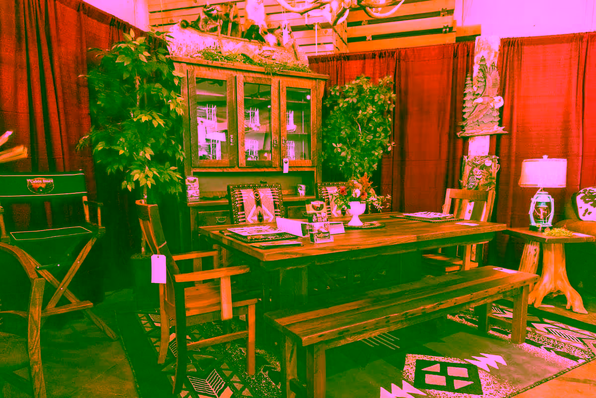 A rustic dining area featuring a wooden table with benches and chairs, surrounded by decorative plants. A glass-fronted cabinet displays various glassware against a red backdrop.