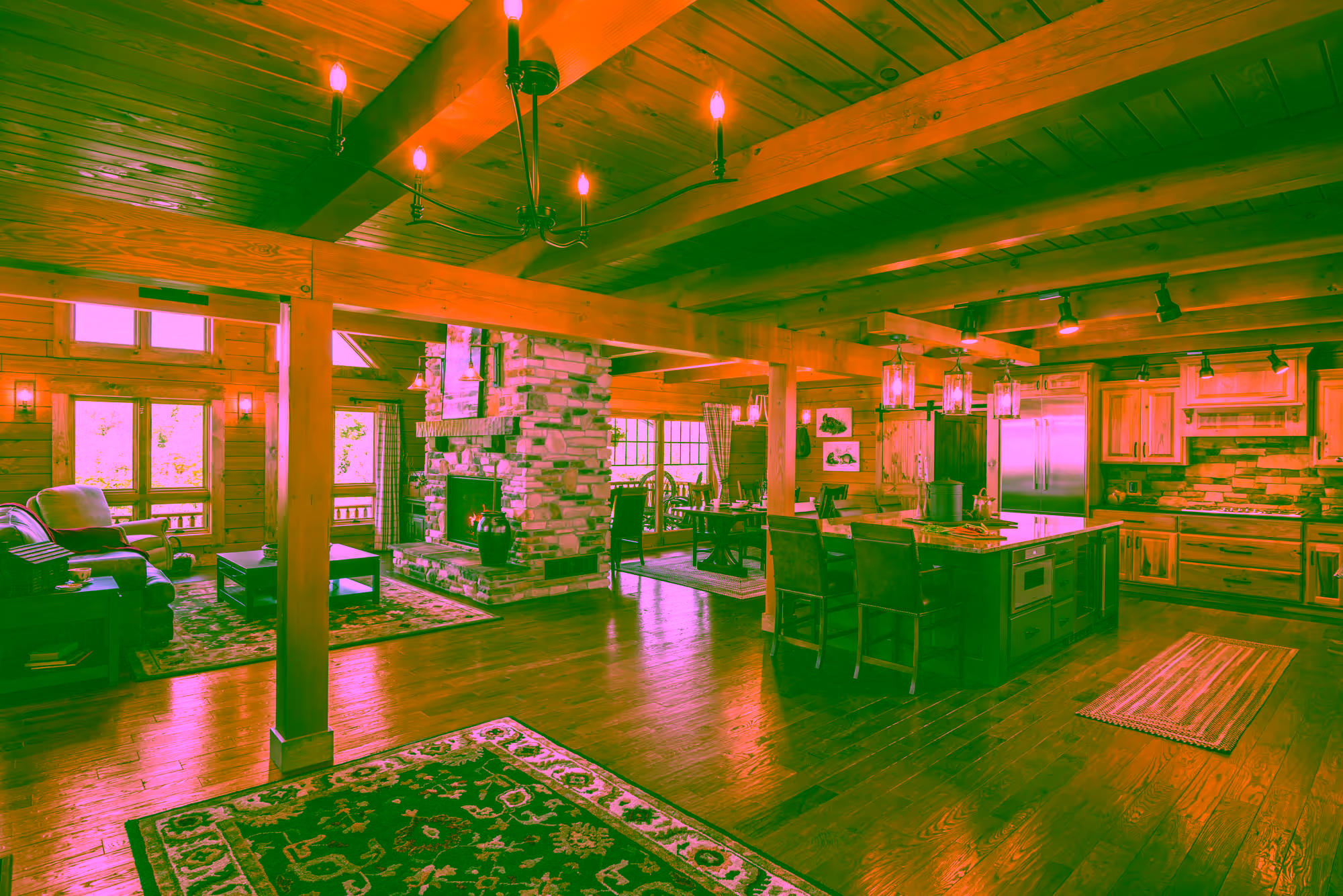 Interior of a spacious log home with wooden beams, featuring a cozy living area with a stone fireplace, and a kitchen island with seating. Large windows provide natural light, and decorative rugs enhance the ambiance.