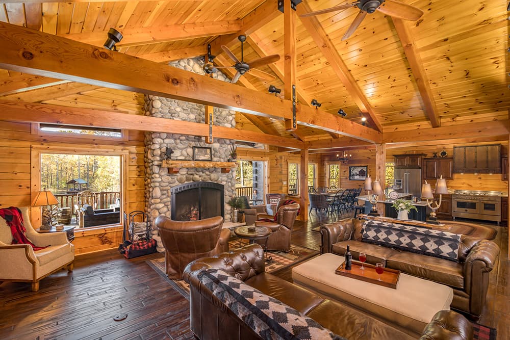 Interior of a spacious log home with wooden beams, featuring a cozy living area with a stone fireplace, and a kitchen island with seating. Large windows provide natural light, and decorative rugs enhance the ambiance.