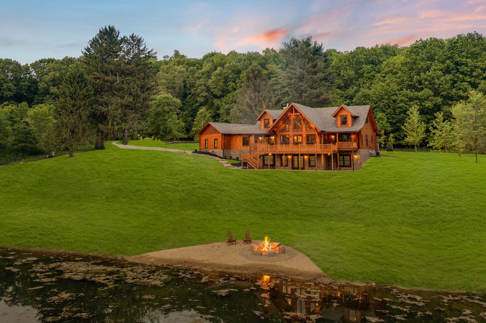 A large log home with multiple gables situated on a grassy hilltop beside a serene pond. A fire pit is visible on the sandy shore in the foreground, surrounded by trees in the background. Ideal for custom homes or log cabins.