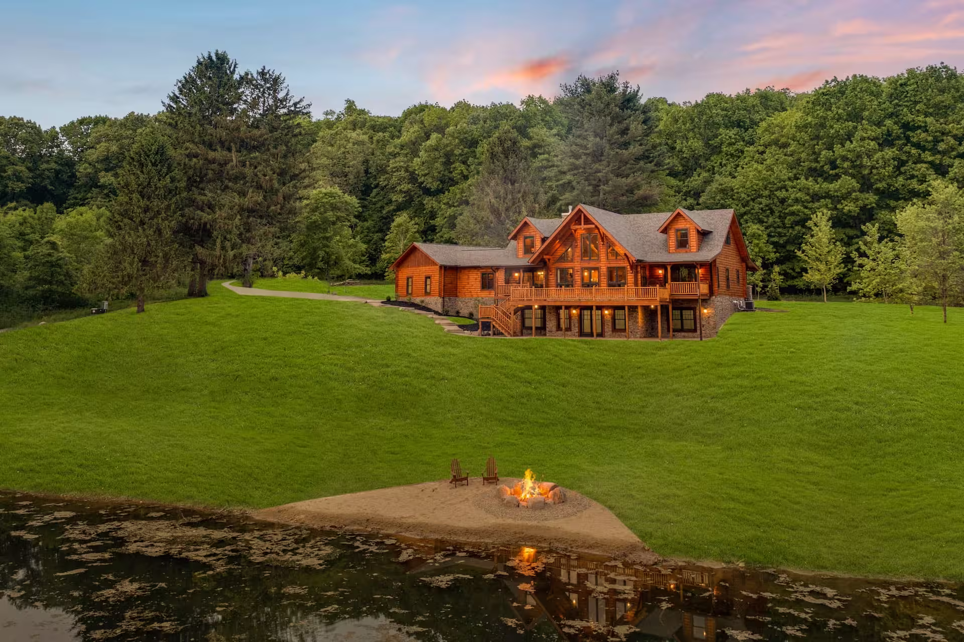 A large log home with multiple gables situated on a grassy hilltop beside a serene pond. A fire pit is visible on the sandy shore in the foreground, surrounded by trees in the background. Ideal for custom homes or log cabins.