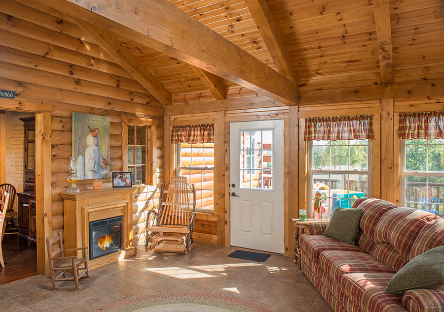 Interior of a log cabin living room featuring wooden beams, a striped couch, a fireplace, and large windows. A rocking chair and a round rug add warmth to the space. The room is well-lit with natural light.