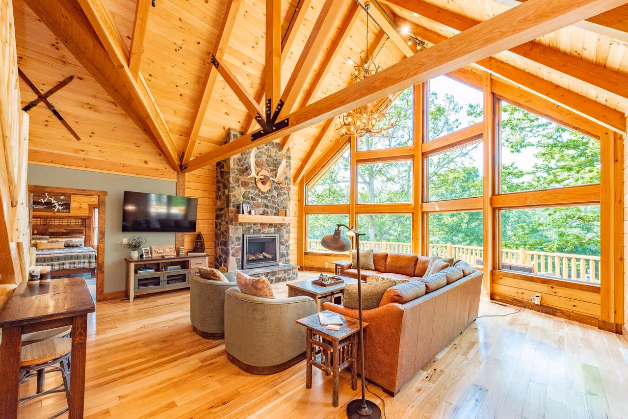 Interior of a log cabin living room featuring wooden beams, a striped couch, a fireplace, and large windows. A rocking chair and a round rug add warmth to the space. The room is well-lit with natural light.