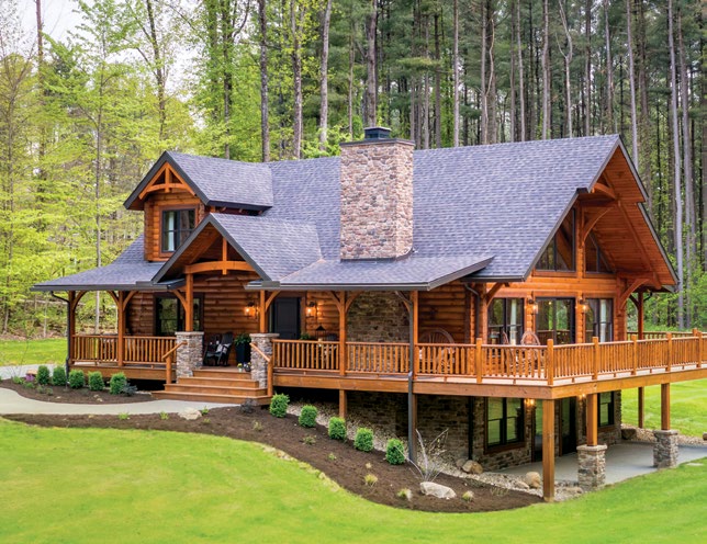 A large log home set on a green hillside next to a calm body of water. The house features multiple gabled roofs and a wooden deck. A small sandy area with a fire pit is visible in the foreground, surrounded by trees.