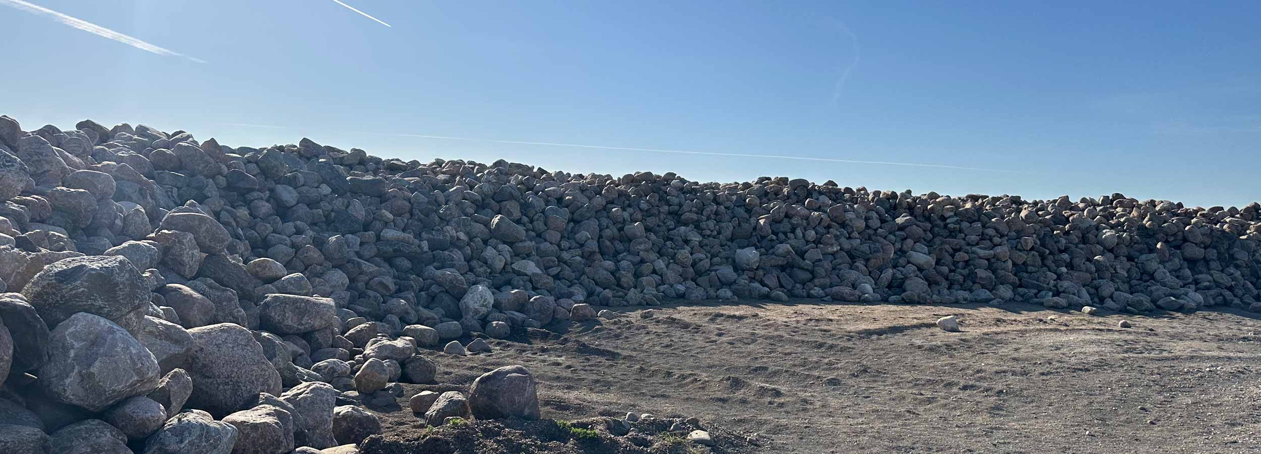 Large pile of rounded gray rip rap stones under a clear blue sky on a dirt ground.