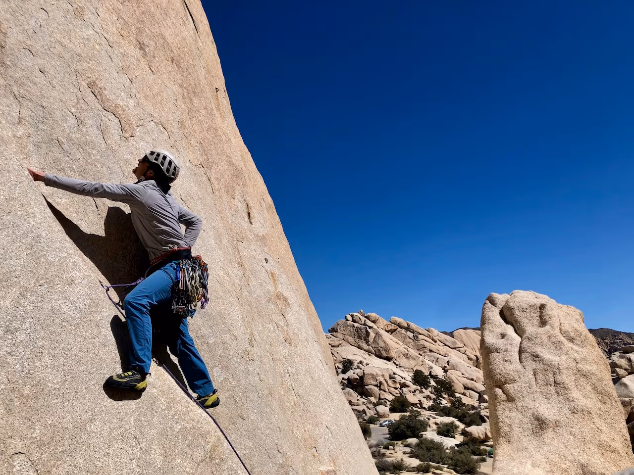A person wearing climbing gear scales a large rock face under a clear blue sky.