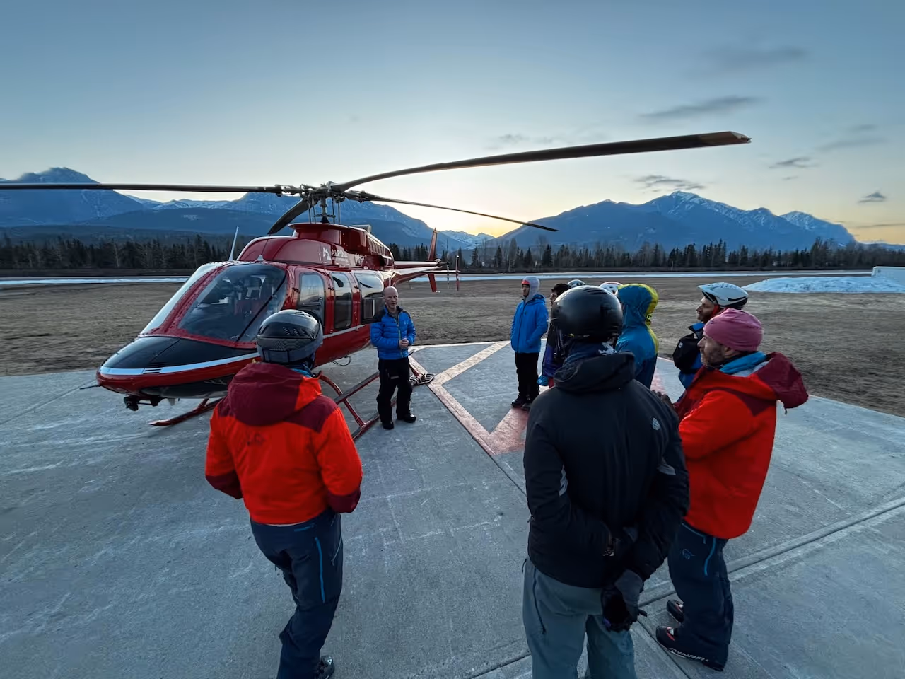 A group of people in helmets listens to a guide near a helicopter on a mountain airfield.