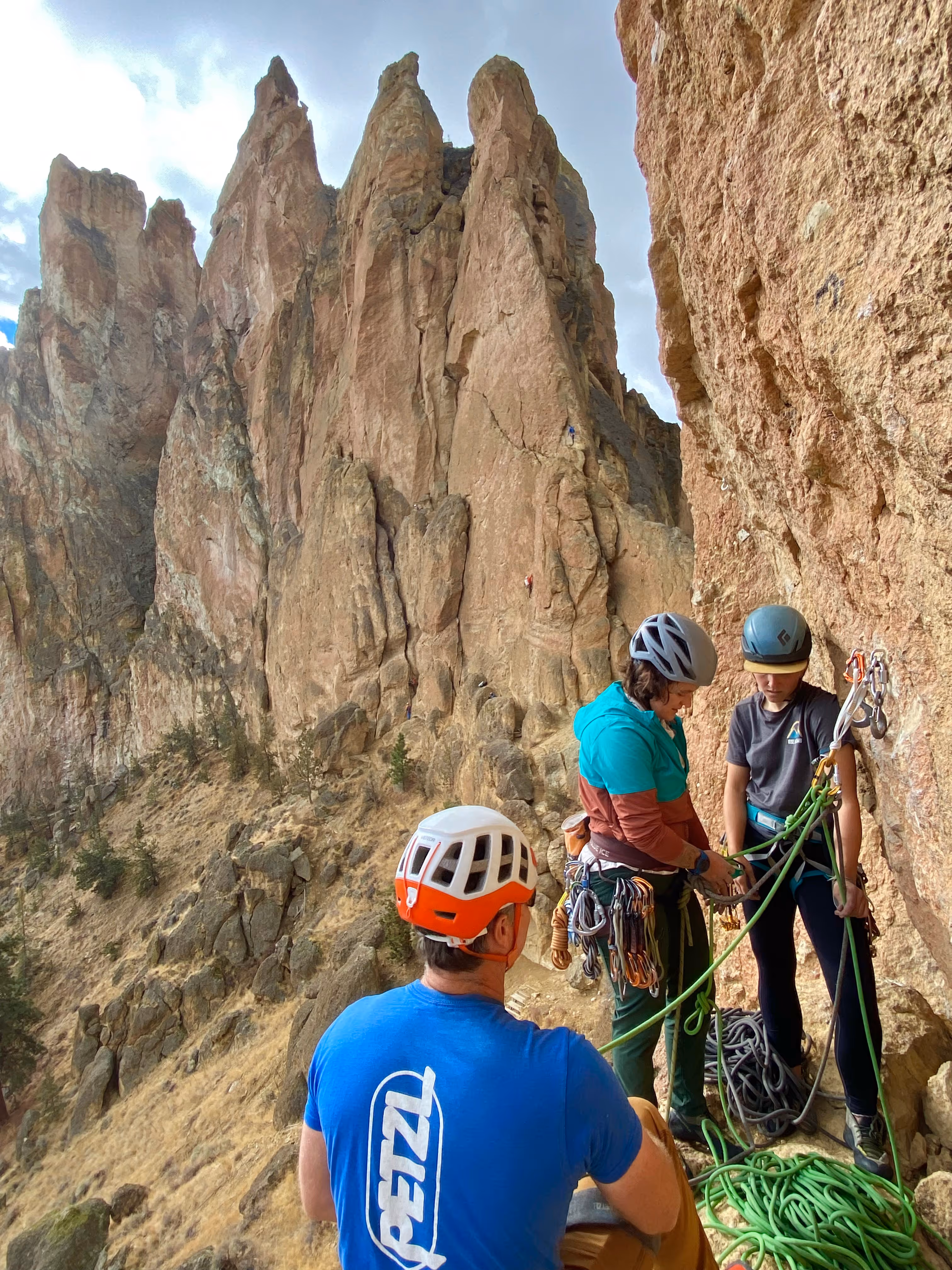 Three people in climbing gear prepare ropes on a rocky ledge near tall, rugged cliffs.