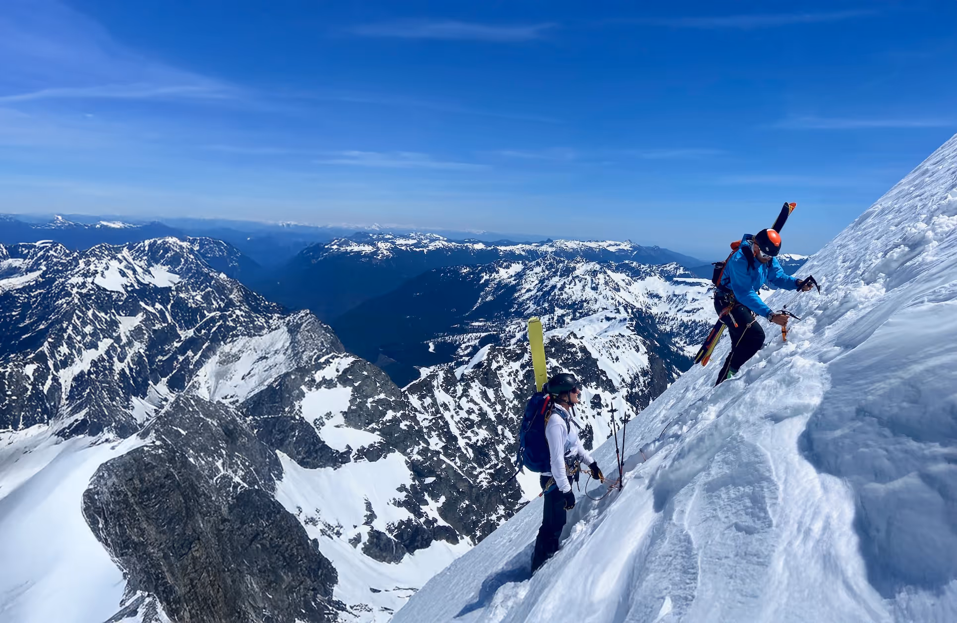 Three climbers wearing snow gear ascend a steep, snowy mountain slope with ski poles.