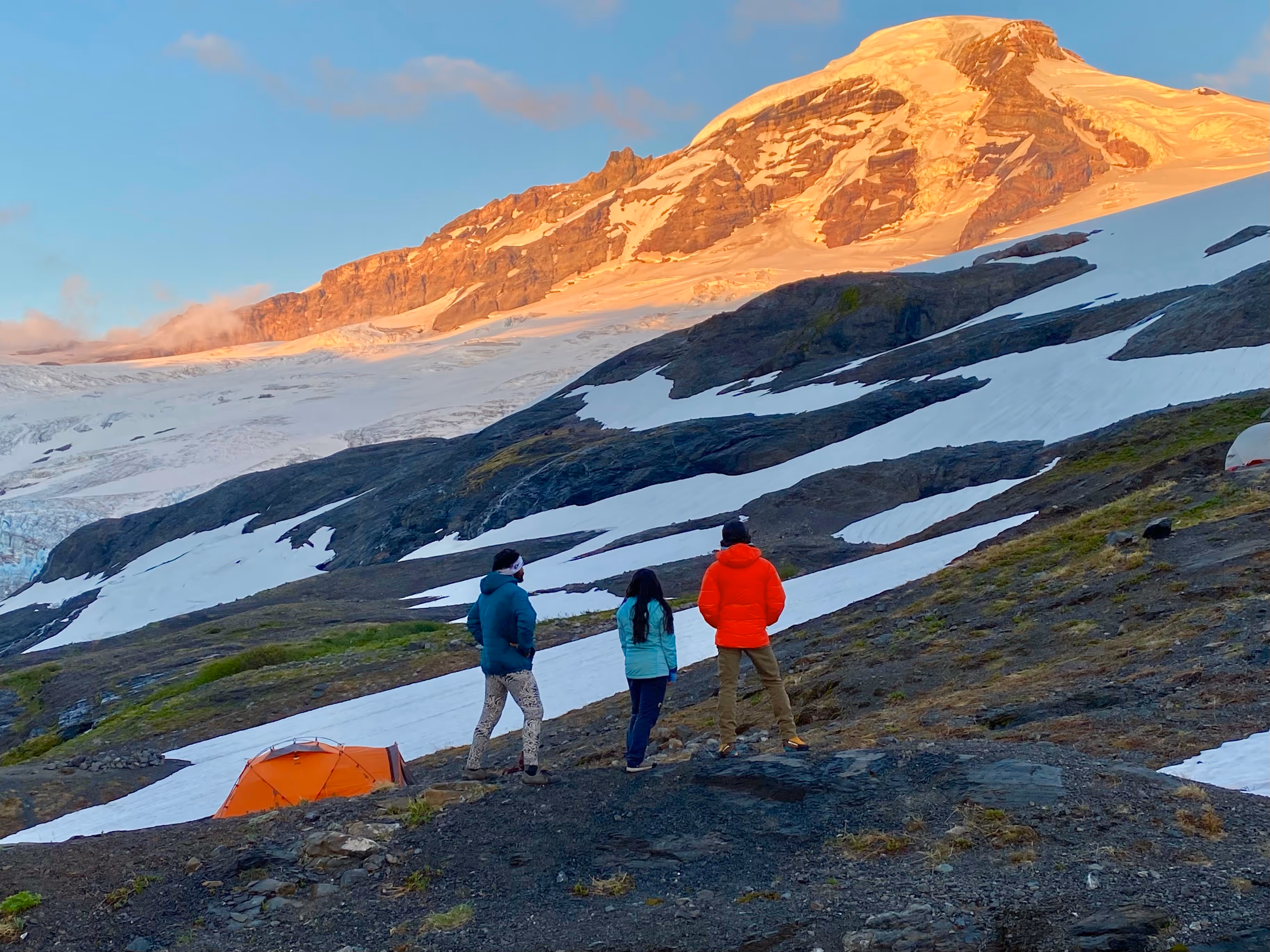 Three people stand near a tent, looking at a snow-covered mountain under a blue sky.