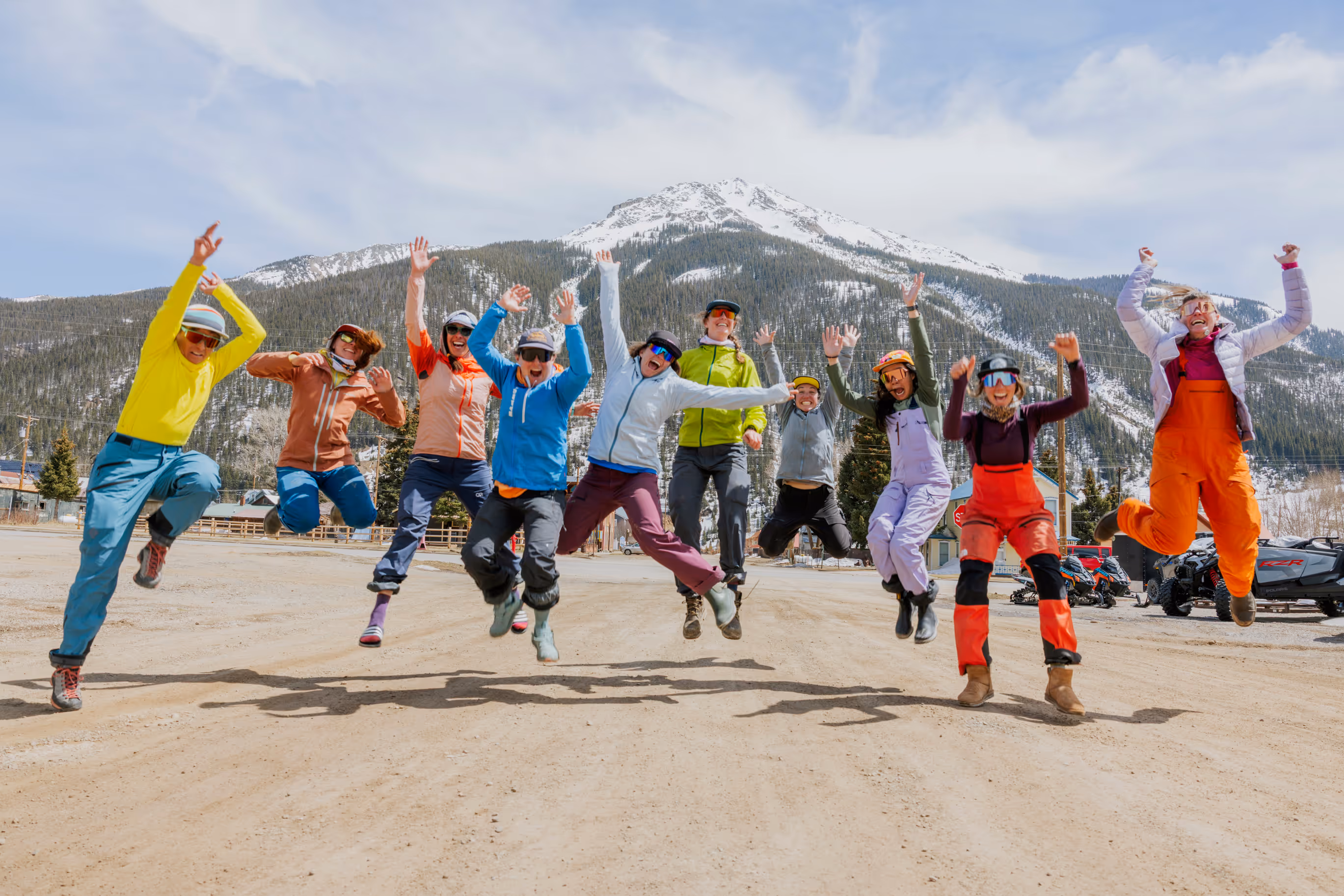 A group of ten people jumping joyfully in a snowy mountain landscape, wearing colorful winter clothes.