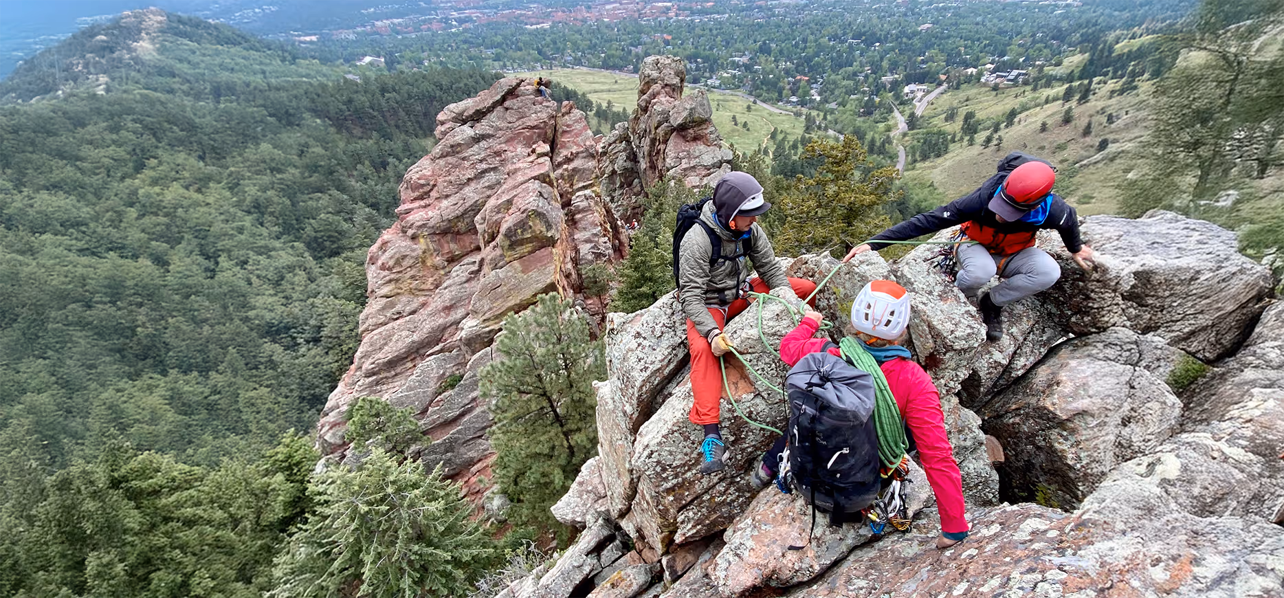 Three climbers, wearing helmets and gear, pause on a rocky peak with a forested view below.
