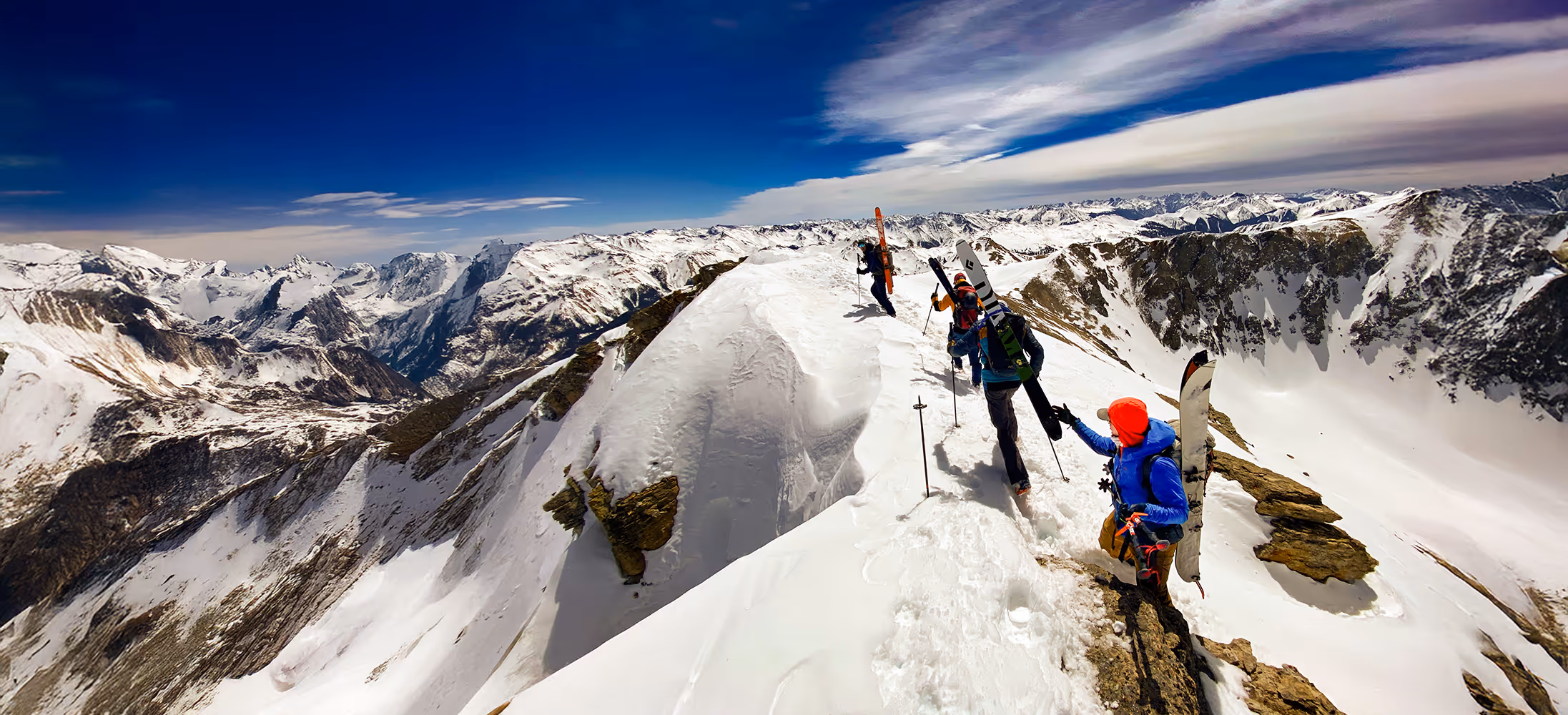 Four people with skis walk along a snowy mountain ridge under a clear blue sky.