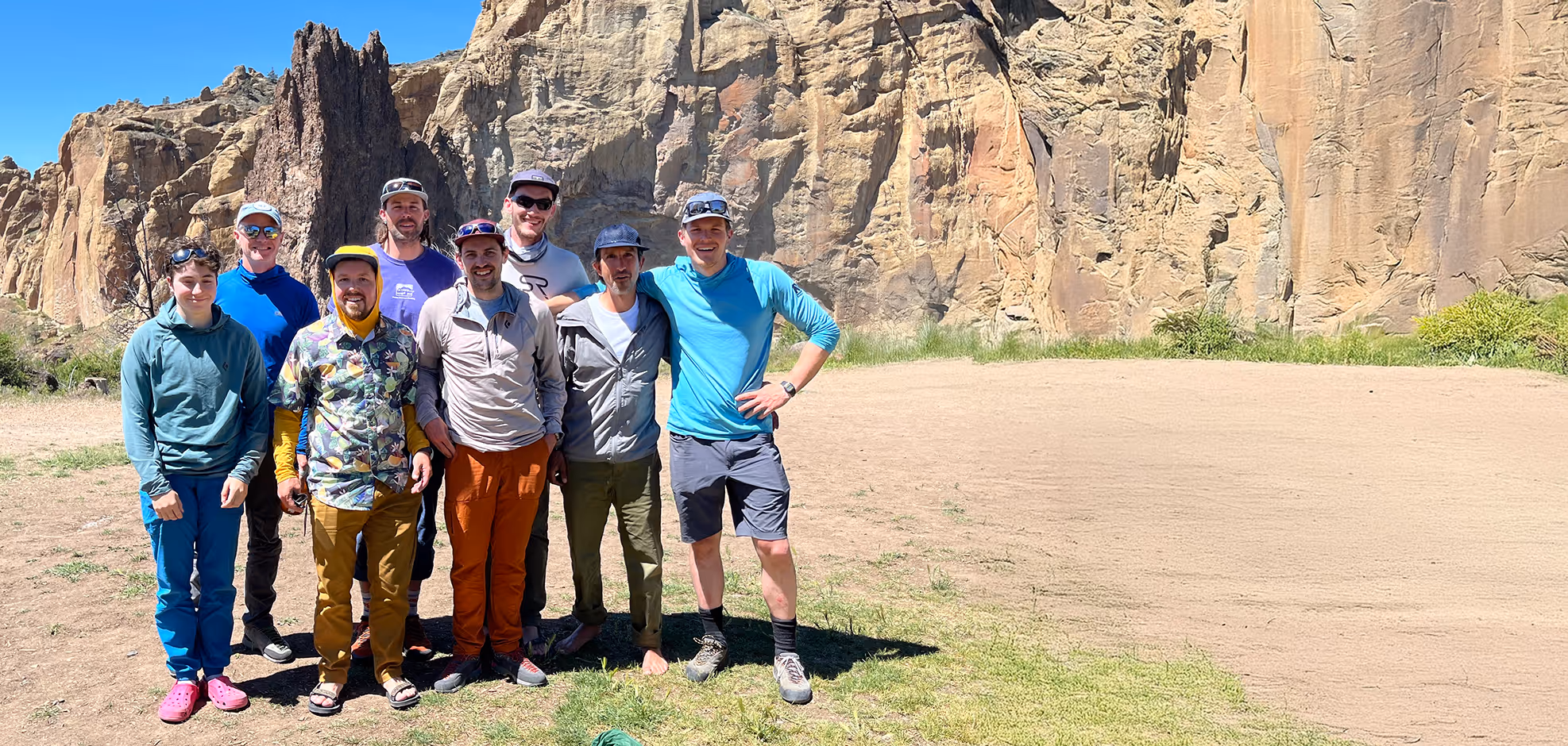 Seven people stand together smiling in front of large rock formations on a sunny day.