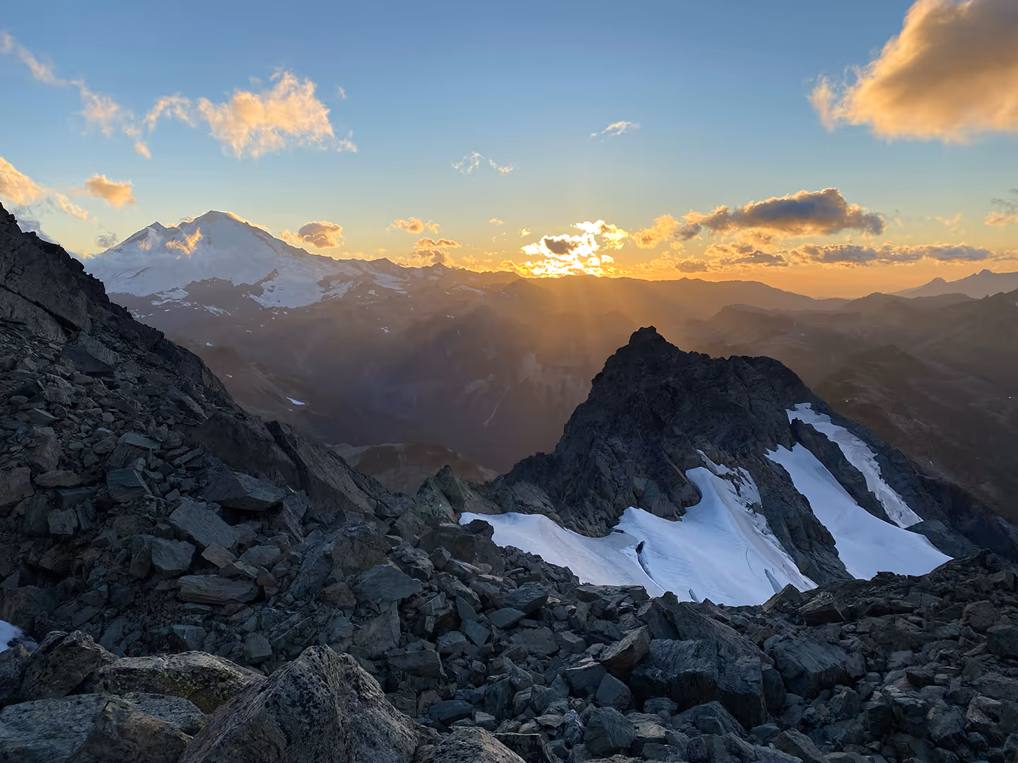 Sunrise over snow-capped mountains with rocky foreground, casting warm light across the landscape.
