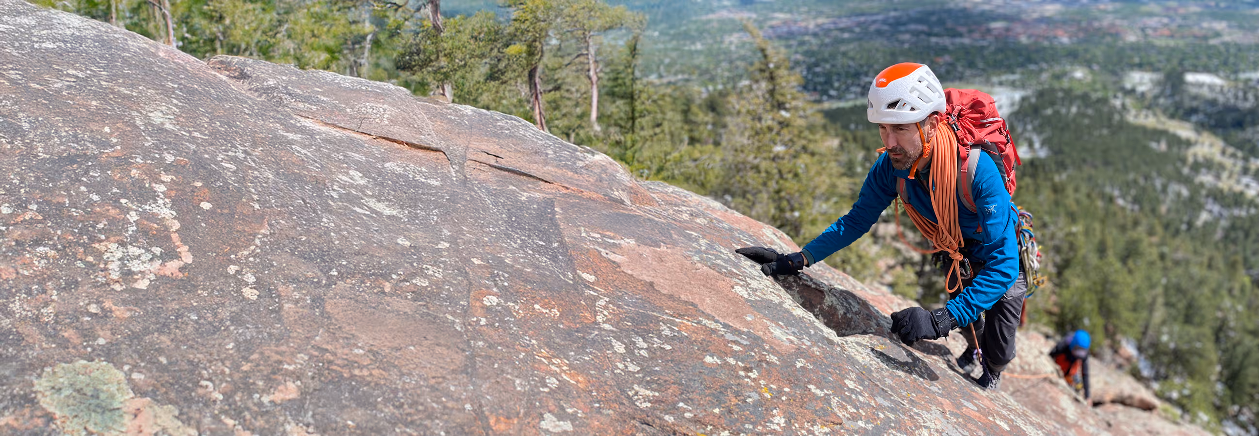 A climber wearing a helmet and backpack ascends a large rock face.