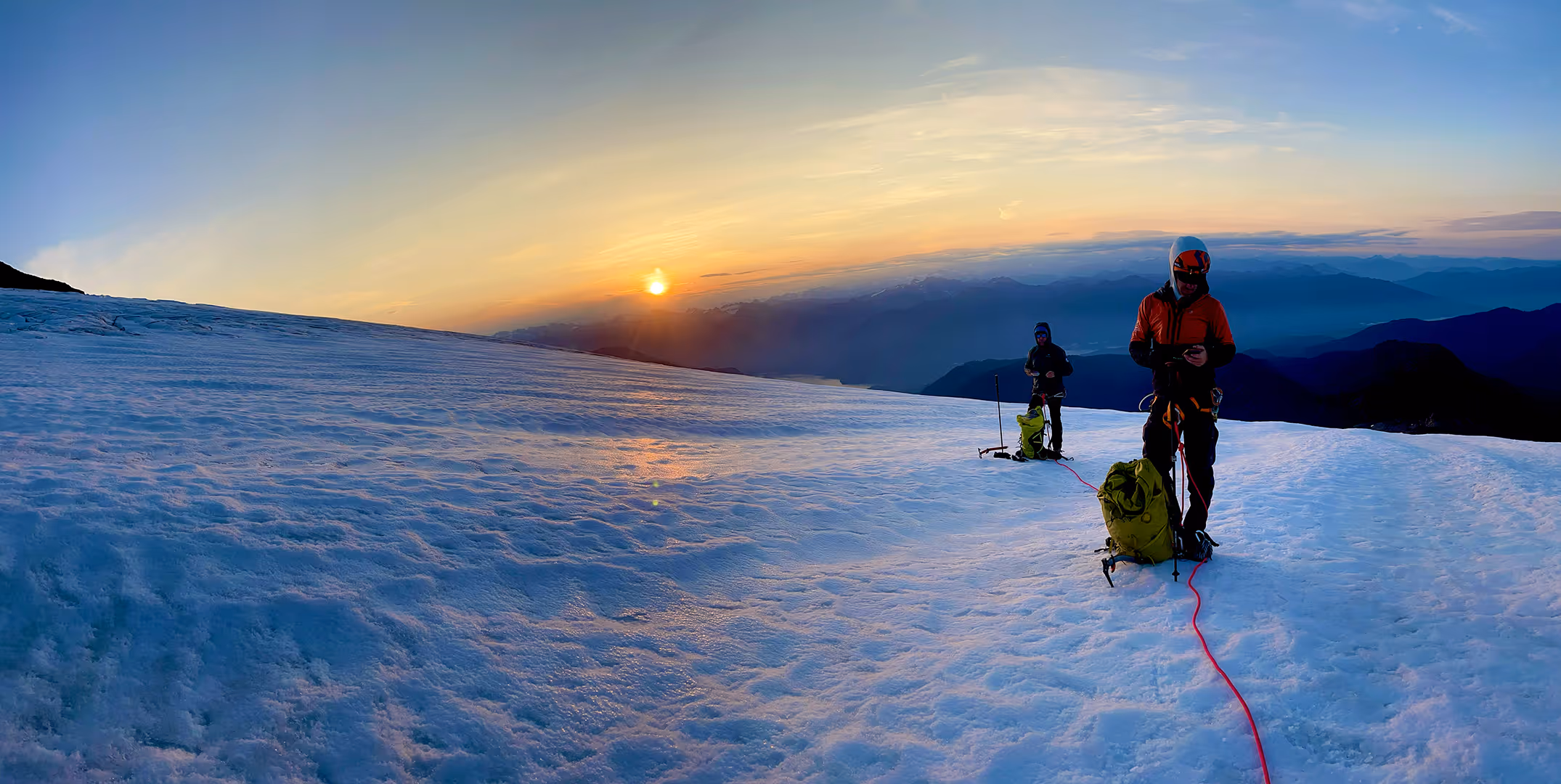 Two climbers stand on a snowy mountain, watching the sunrise with gear and ropes.