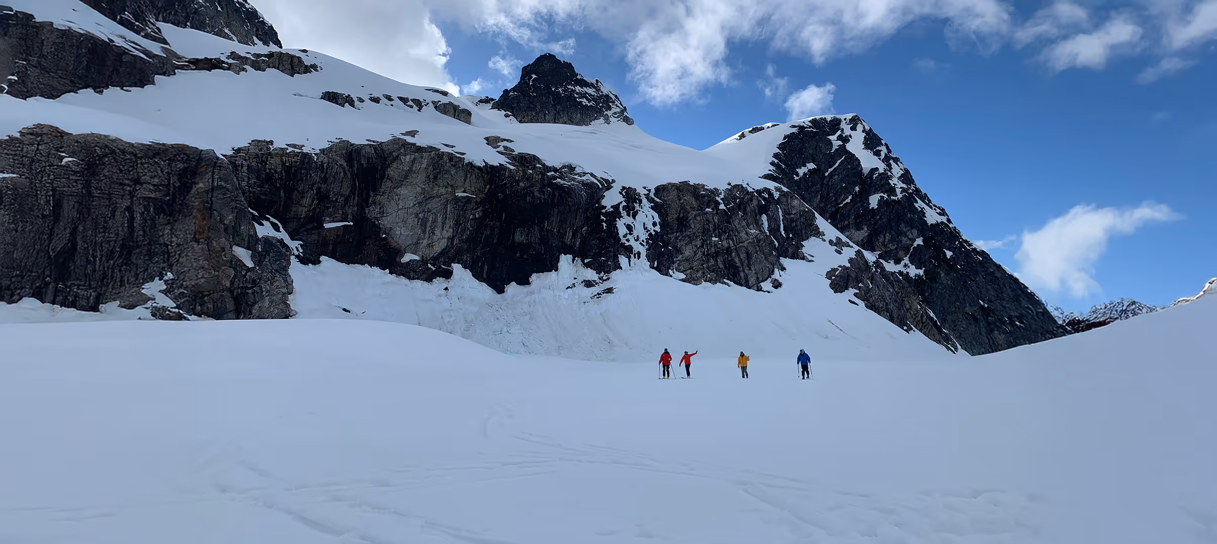 Four people are walking across a snowy field near large, rocky mountains under a blue sky.