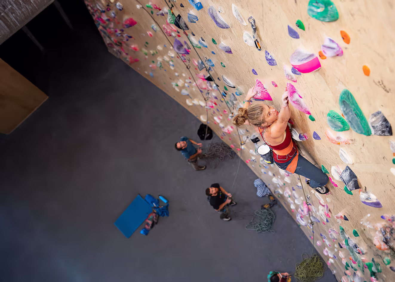 A woman climbs an indoor rock wall while people watch from below.
