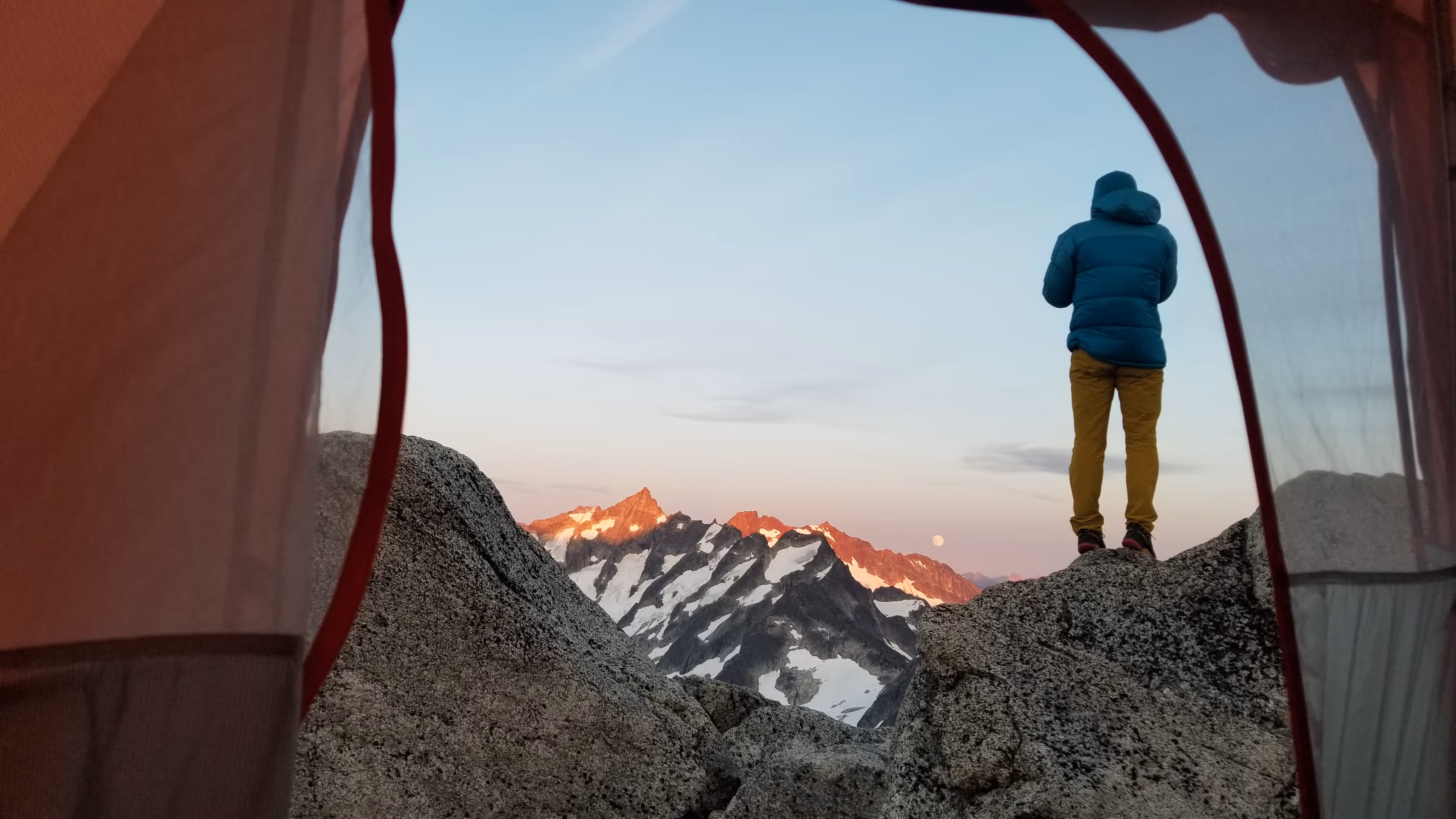 Person in a blue jacket stands on rocks, looking at snow-capped mountains during sunrise.