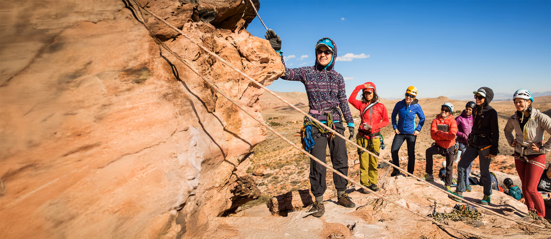 A group of climbers on a rocky ledge listens to a person explaining climbing techniques.