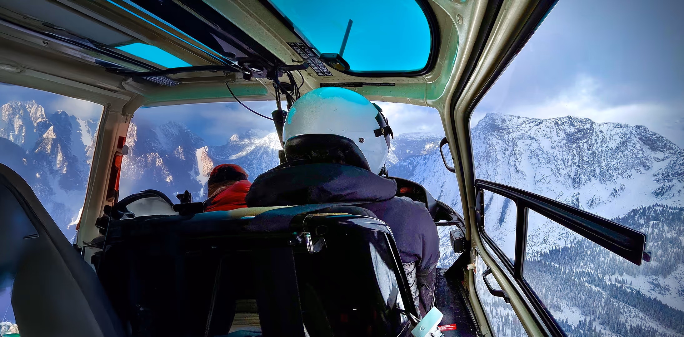 Two people inside a helicopter fly over snowy mountains, with the pilot wearing a white helmet.
