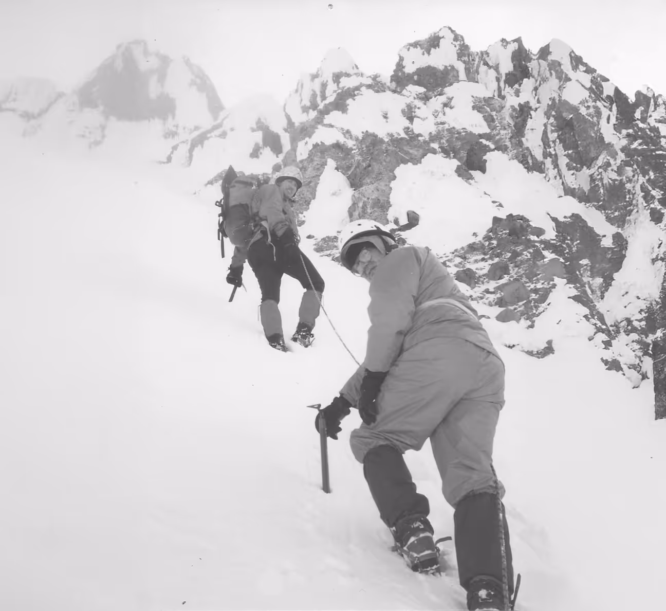Two climbers in winter gear ascend a snowy mountain slope using ice axes for support.