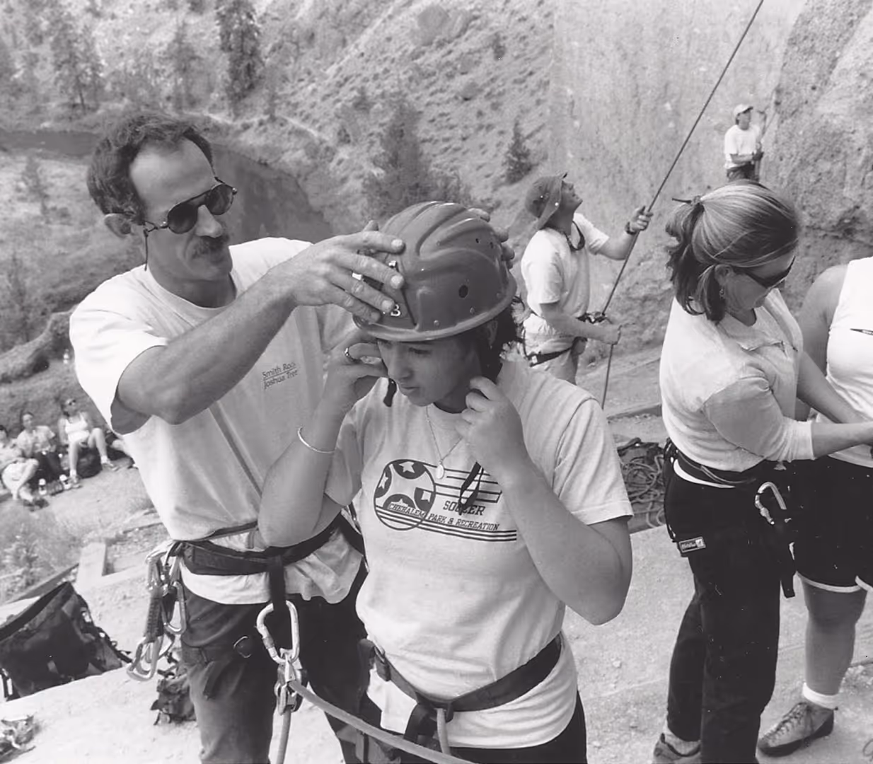 A man helps a young woman adjust her helmet before climbing. Others prepare in the background.