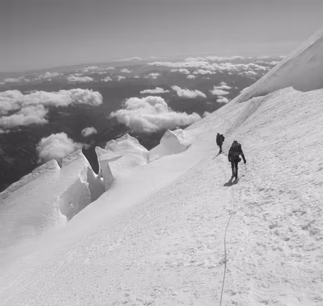 Two climbers in gear navigate a snowy mountain slope under a cloudy sky.
