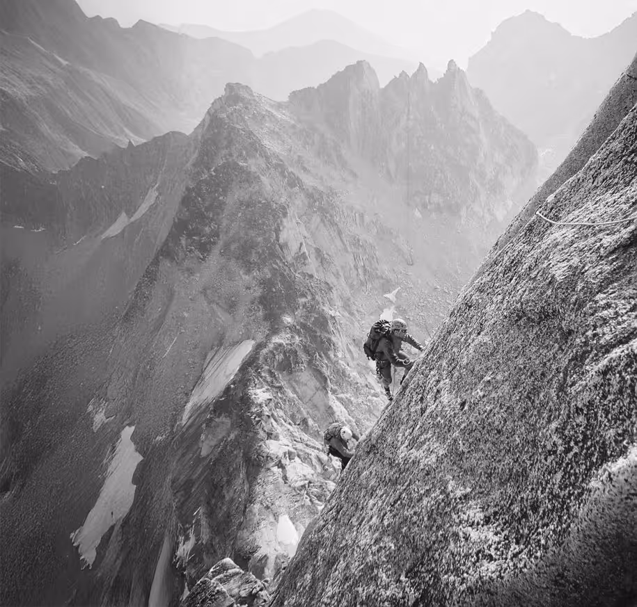 Two climbers ascend a steep, rocky mountain slope with rugged peaks in the background.