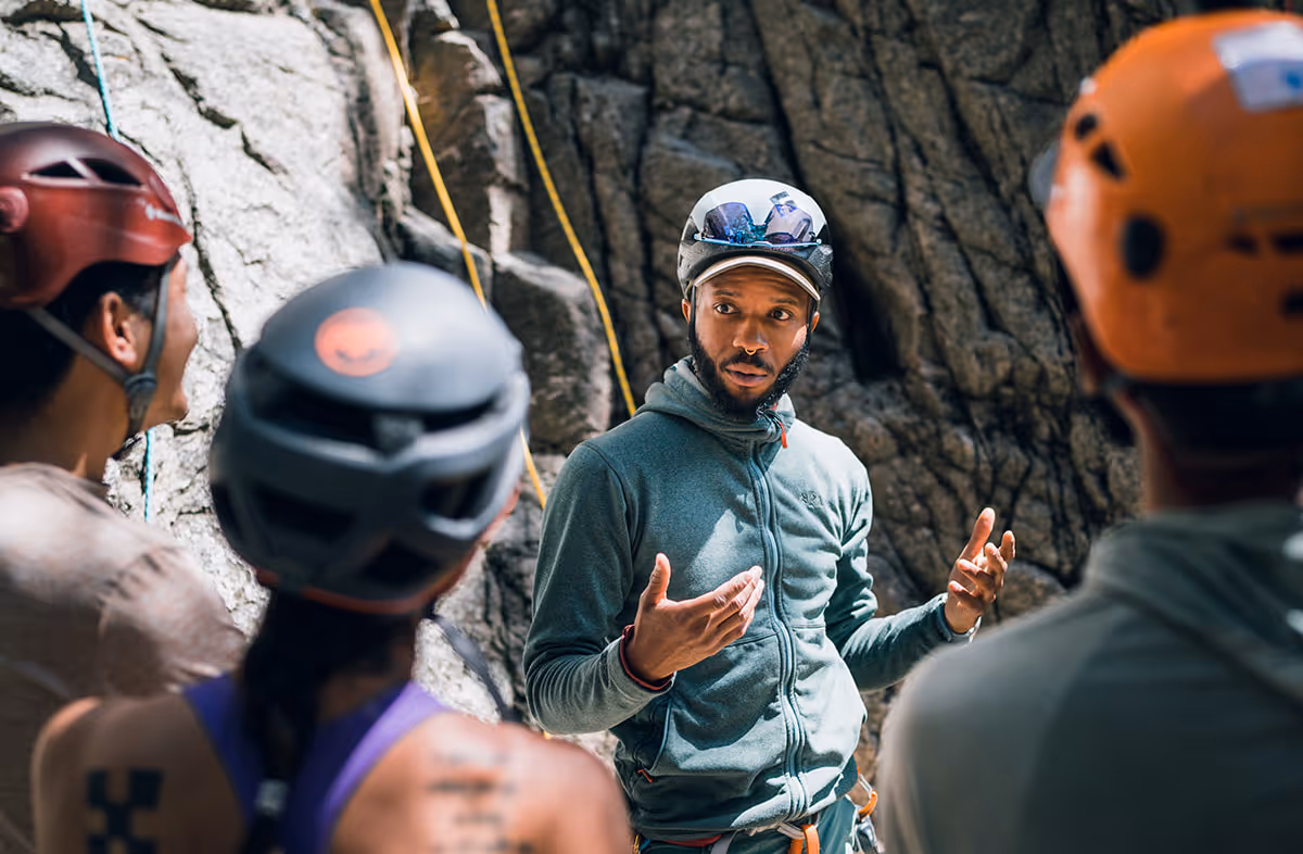 A person in climbing gear instructs a group near a rock wall.