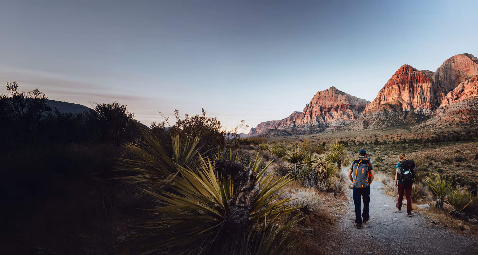 Two people with backpacks walk on a trail towards large red rocky mountains at sunset.