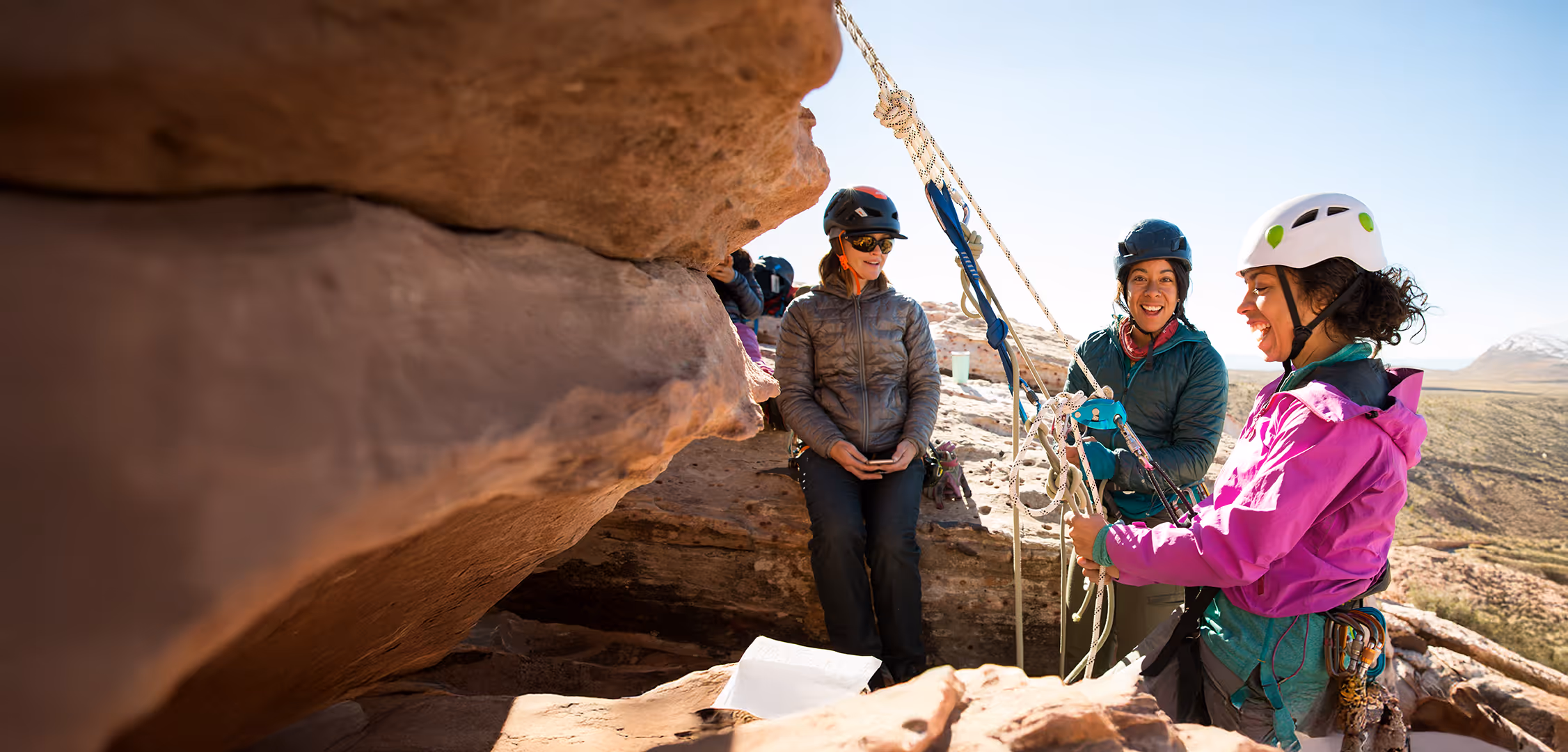 Three people with helmets prepare climbing equipment at a rock formation, smiling and chatting.