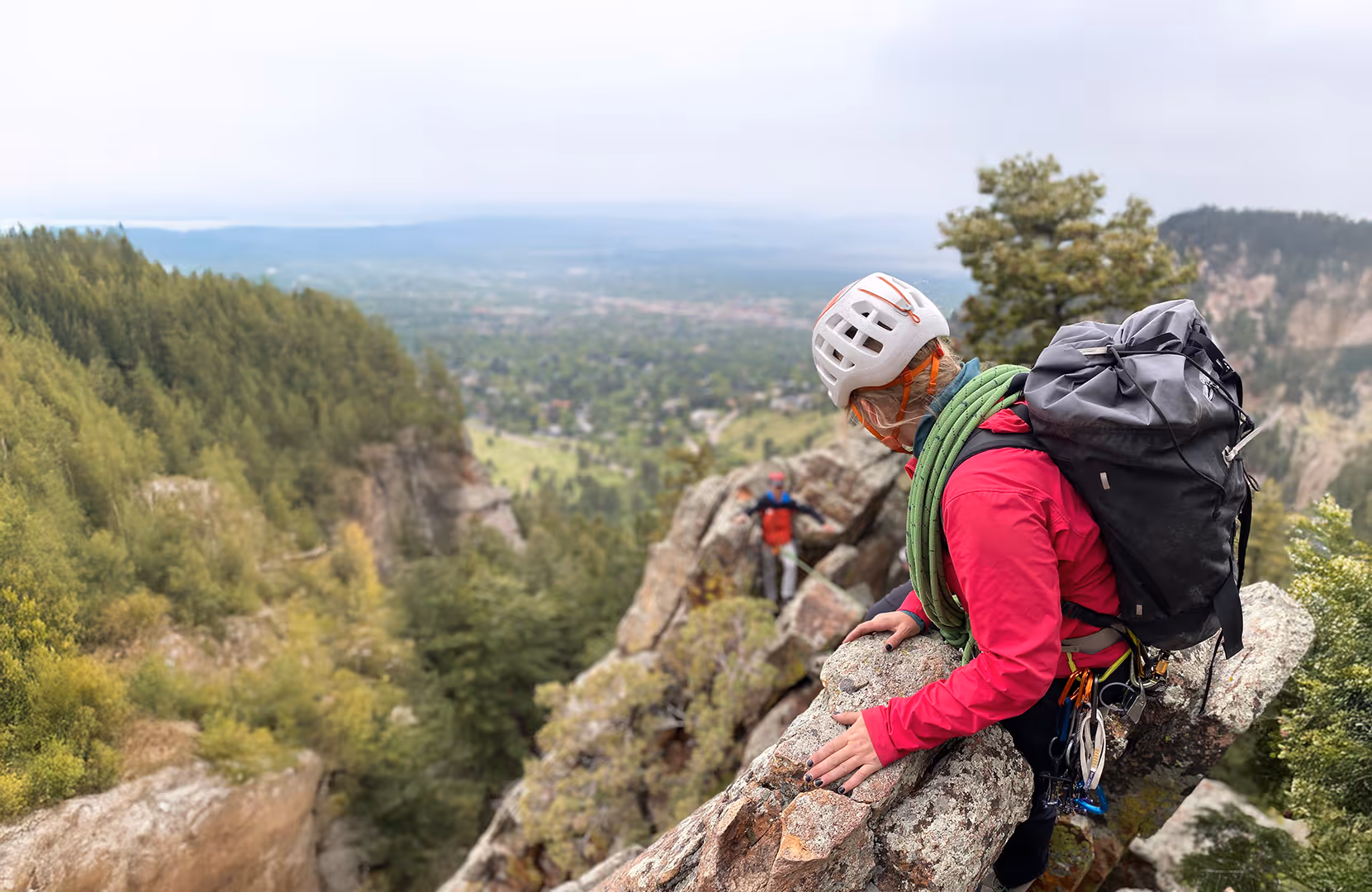 A climber wearing a helmet and backpack climbs a rocky ridge, with a forested valley below.